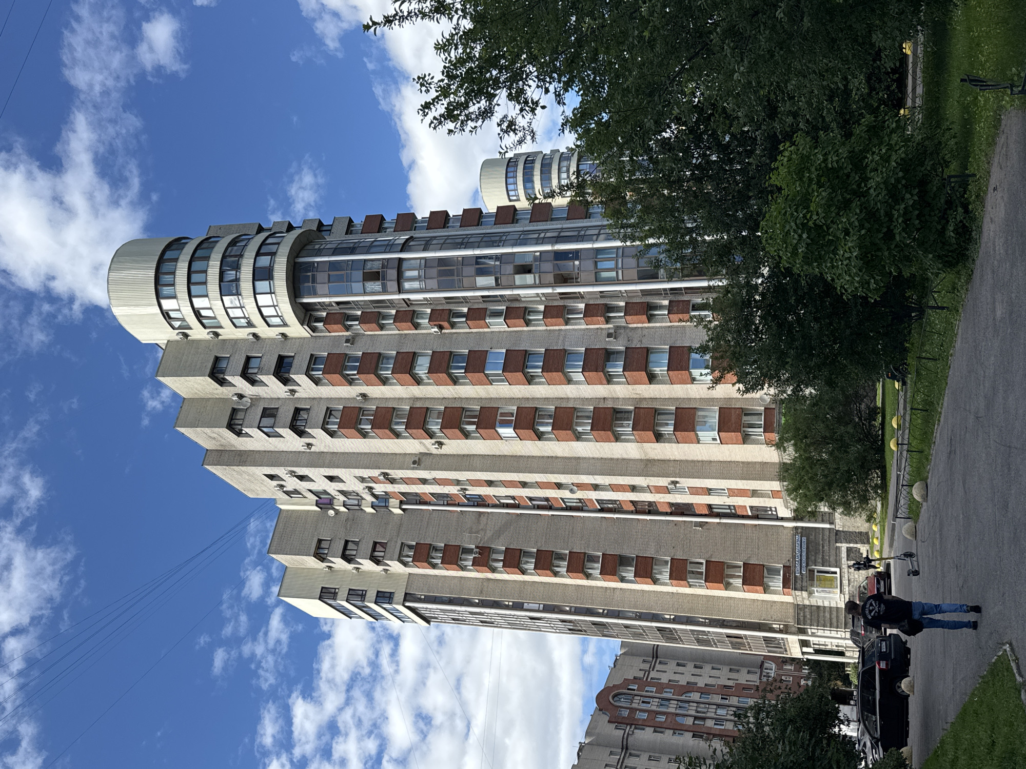 This image depicts a residential building in Saint Petersburg, Russia, likely on a sunny day with scattered clouds. The building is tall, modern, and constructed with light-colored bricks and terracotta accents. It features numerous windows and balconies, and a distinctive curved section with a cylindrical top on the right side. The foreground shows a paved pathway with grassy areas and trees on either side. A man in a black t-shirt and blue jeans is walking away from the camera down the pathway. To the left, several parked cars are visible, including a black SUV and a red sedan. A child's scooter stands upright near the man. The street is bordered by round, light-colored bollards. In the background, other residential buildings and lush green trees are present, indicating a suburban or city park setting. The overall impression is one of a well-maintained residential area under a clear blue sky.