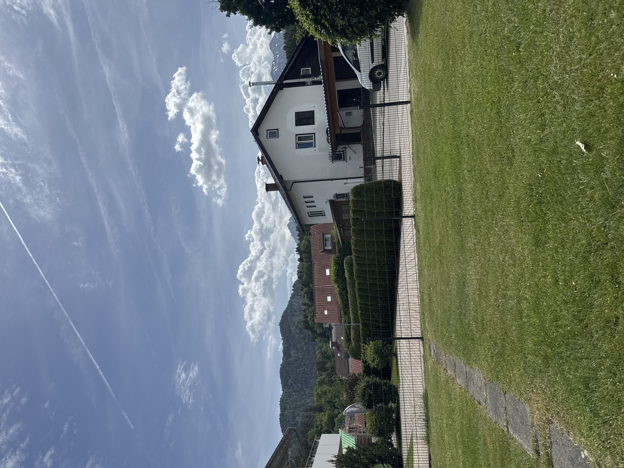 The image shows a residential area in Füssen, Germany. A white house with a dark brown roof is the most prominent feature, situated behind a dark green hedge and a black chain-link fence. A white van is parked near the house. Behind the house, there is a view of trees, rooftops of other houses, and a mountain. The sky is blue with streaks and cumulus clouds. A contrail crosses the upper left portion of the sky. A green lawn is visible on the right side of the image, bordered by a paved walkway.