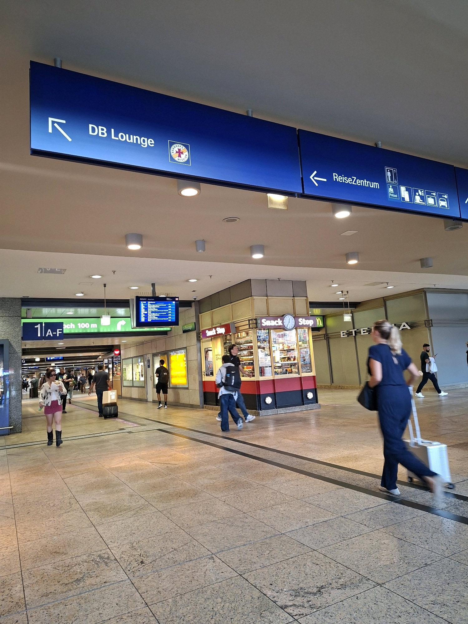 The image depicts a busy indoor environment, likely a train station, with a focus on directional signage and pedestrian traffic. In the upper part of the image, a large blue sign indicates "DB Lounge" with an arrow pointing left, and another sign to the right reads "ReiseZentrum" with an arrow pointing left and various pictograms representing services like toilets, elevators, police, luggage, and parking.
Below the main signs, a green directional sign displays "1 | A-F" and "nach 100 m" (to 100 m), suggesting a platform or section designation. Further down, a digital display board shows train departure information.
Several individuals are visible navigating the space. In the foreground right, a woman in a dark blue jumpsuit is pulling a silver wheeled suitcase, her movement blurred by motion. To the left, a person in a light-colored top and a red skirt is walking, also with a blurred effect. Other pedestrians, some carrying bags or suitcases, are scattered throughout the scene, moving in various directions.
A "Snack Stop" kiosk with bright lighting and stocked shelves is visible in the mid-ground. In the background, the architecture suggests a modern commercial or transit hub with tiled flooring, overhead lighting, and architectural elements. The overall atmosphere is one of transit and movement within a public space. Based on the "DB Lounge" and "ReiseZentrum" signage, the location is identified as Cologne Hauptbahnhof (main station) in Cologne, Germany.