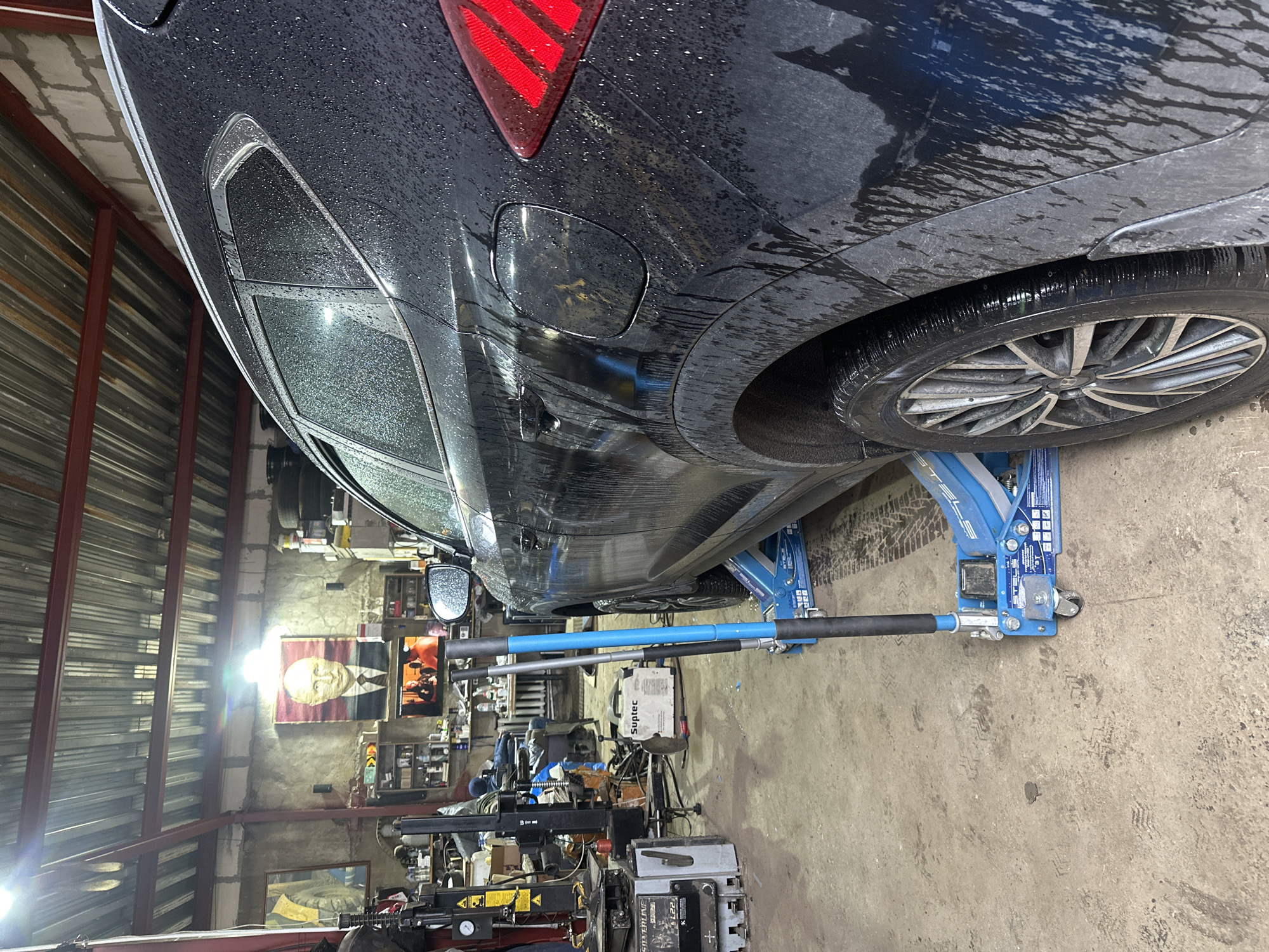 The image shows a black car raised on a blue hydraulic jack in what appears to be a garage or workshop. The car is wet, suggesting it may have recently been washed or driven in the rain. The rear driver-side wheel is visible, as is a portion of the taillight and fuel filler door. The garage interior is cluttered, with various tools, equipment, and shelves visible in the background. A portrait of Lenin hangs on the wall.
The location, Dubna, Russia, is inferred from the portrait of Lenin, which is often found in former Soviet territories. The lack of more distinct identifying markers necessitates reliance on this cultural artifact.