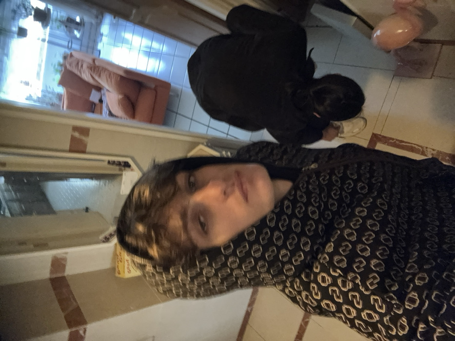 The scene depicts an interior residential setting, specifically an entryway or hallway, located in the city of Vincennes, France.
In the foreground, a young adult male with brown hair is visible from the chest up, directly facing the camera with a neutral expression. He is wearing a dark hooded sweatshirt patterned with repeating light-colored stylized 'N' or oval shapes.
To his right and slightly behind, a second individual is positioned with their back to the camera, bent forward at the waist. This individual wears dark clothing, possibly a black hoodie and pants, and white athletic shoes. Their dark hair is visible, appearing to be pulled back. The individual's action suggests they are looking at or interacting with something on the floor.
The floor throughout the visible area is composed of light-colored square tiles with reddish-brown linear borders. The walls are light-colored, featuring horizontal reddish-brown stripe accents. To the left, a doorway opens into another room with visible cabinetry and tiled flooring. To the right, a wider opening leads into a brightly lit room featuring a large, orange, L-shaped sectional sofa. This room also has tiled flooring and visible plants near a window or glass door. A deflated pink balloon lies on the floor in the lower right portion of the frame, near the second individual. A partially obscured yellow box, displaying text that begins with "GOOD GR...", is visible on a surface behind the first individual.