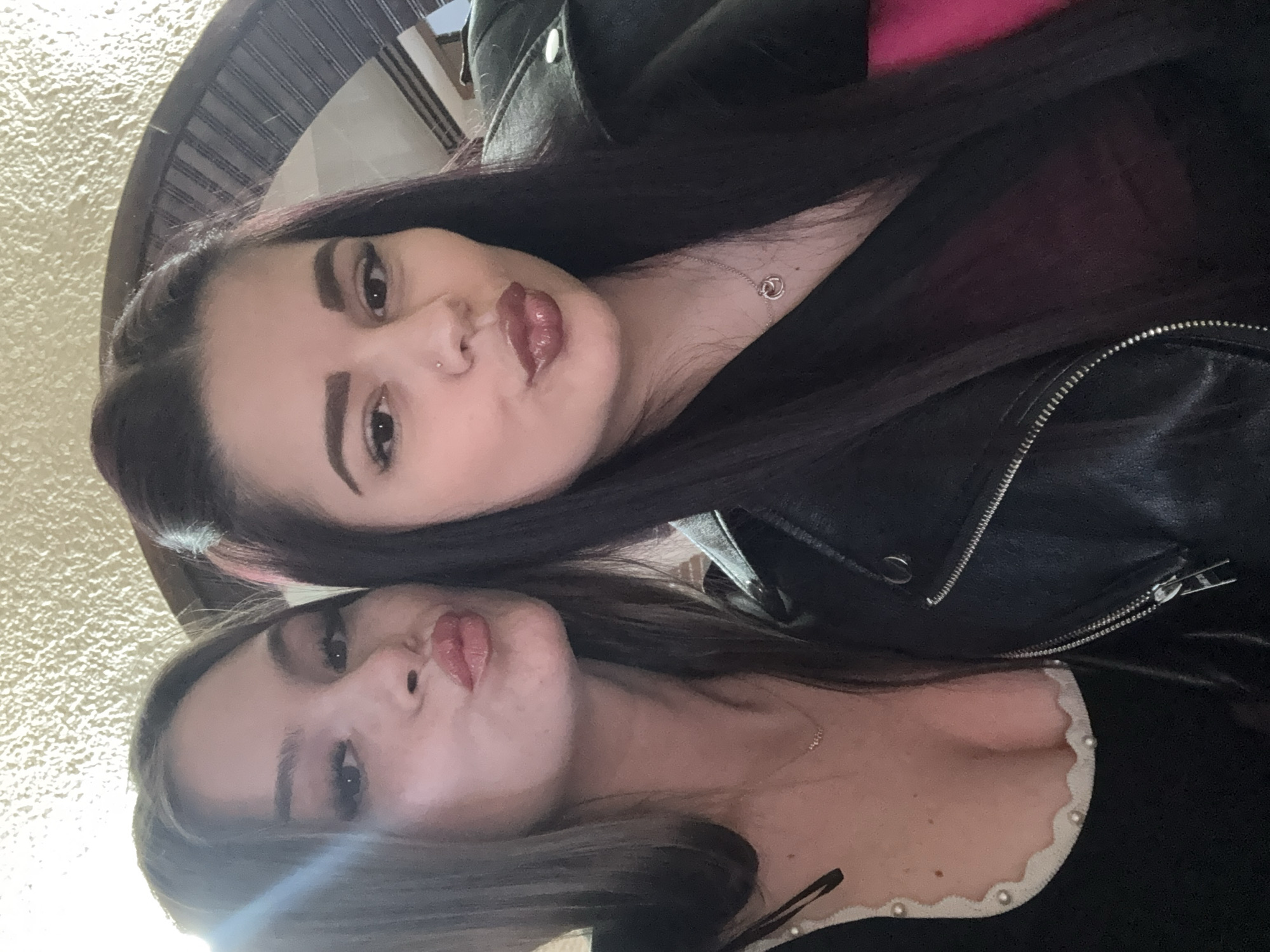 Two young women are posing closely together, looking at the camera with puckered lips as if giving a kiss. The woman at the top of the frame has dark, long hair, a nose piercing, and is wearing a black leather jacket. The woman at the bottom of the frame has light brown hair and is wearing a black top with a white, scalloped neckline. The background is out of focus, but appears to be an interior setting with a textured wall and part of a dark, decorative object. The image does not indicate the location of Parvomay, Bulgaria.