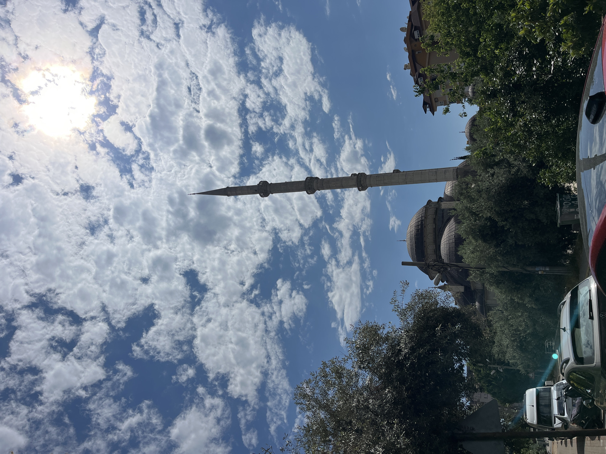 The image shows a view of a mosque, likely in Alanya, Turkey, judging by the architecture of the mosque and surrounding buildings. The minaret of the mosque is visible stretching high into the sky, which is filled with scattered white clouds and a bright sun. The dome of the mosque is also visible, partially obscured by trees. Surrounding the mosque are buildings and foliage, including trees and greenery that indicate a residential or urban area. Vehicles are visible on a street, suggesting the mosque is located in an accessible area. The overall impression is of a sunny day with a view towards the mosque, showing the integration of religious architecture into the cityscape of Alanya.