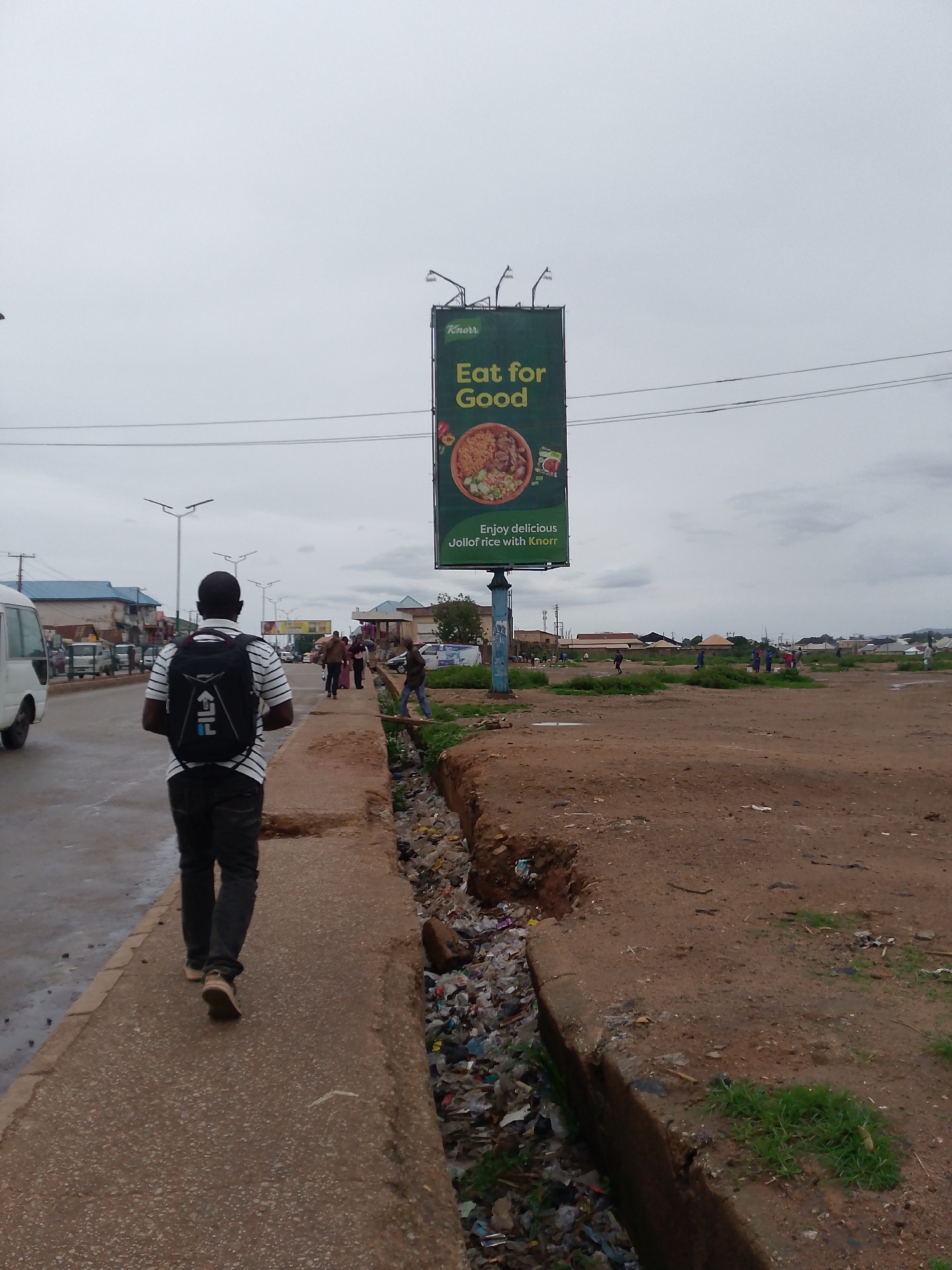 This outdoor photograph captures a street scene in Bukuru, Nigeria, on an overcast day, indicated by the uniformly light grey sky and the wet appearance of the road. In the foreground, a man wearing a striped short-sleeve shirt, dark trousers, and light-colored shoes walks away from the camera on a concrete sidewalk. He carries a black backpack with a prominent "FILA" logo.
To the man's right, a wide, open drainage ditch runs parallel to the sidewalk, filled with a considerable amount of accumulated trash and debris, including plastic wrappers and other refuse. Beyond this ditch lies a large, unpaved area of reddish-brown dirt, with sparse patches of green grass.
A prominent green billboard stands in the mid-ground, featuring the "Knorr" logo at the top. Its main slogan reads "Eat for Good," and it displays an appetizing image of a bowl of Jollof rice, accompanied by the text "Enjoy delicious Jollof rice with Knorr."
Further down the road, other pedestrians are visible walking on the sidewalk. A white minibus is partially seen on the far left, suggesting public transportation. Utility poles, streetlights, and various buildings, some with blue roofs, line the road in the distance. Several individuals are also scattered across the distant dirt field on the right. The scene depicts a busy, semi-urban environment with ongoing daily life and commercial advertising.