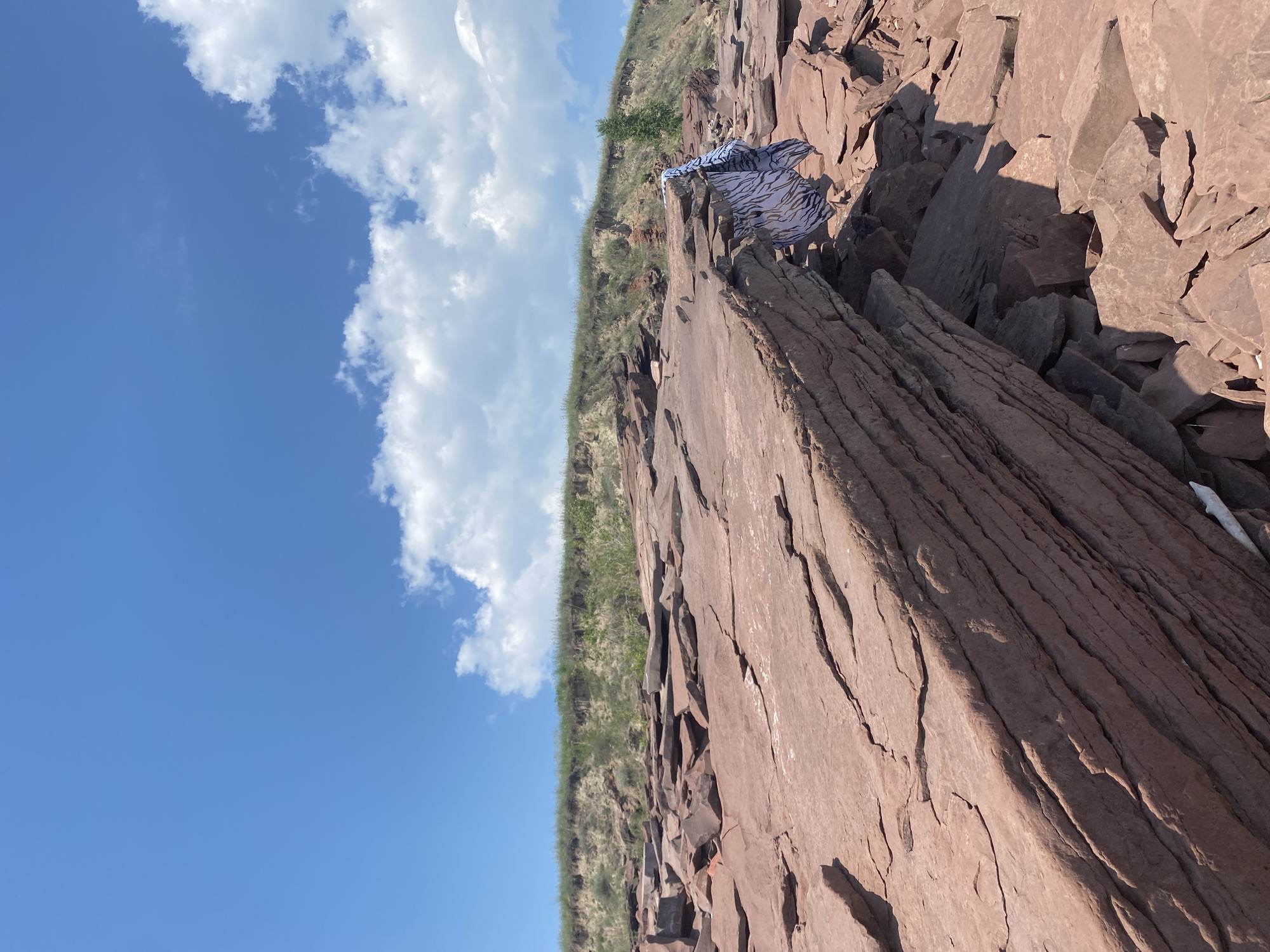 A wide shot captures a landscape of flat, reddish-brown rocks under a bright blue sky dotted with fluffy white clouds. In the background, a grassy embankment slopes upwards, topped with more vegetation. A patterned blue and white garment is draped over some of the rocks, near the right side of the frame.