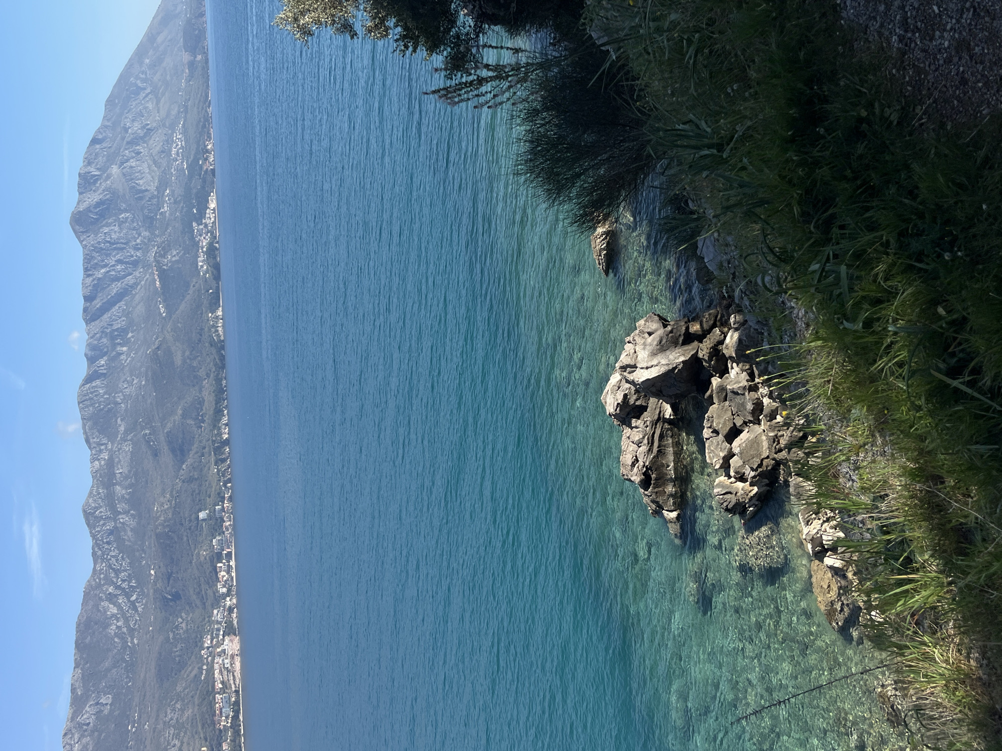 The image shows a view of the Adriatic Sea, likely taken near Budva, Montenegro. The water is clear and turquoise, with rocks and vegetation lining the shoreline. On the left, a mountain range is visible, with a town built along its base. The scene appears peaceful and sunny. There are no people visible in the image.