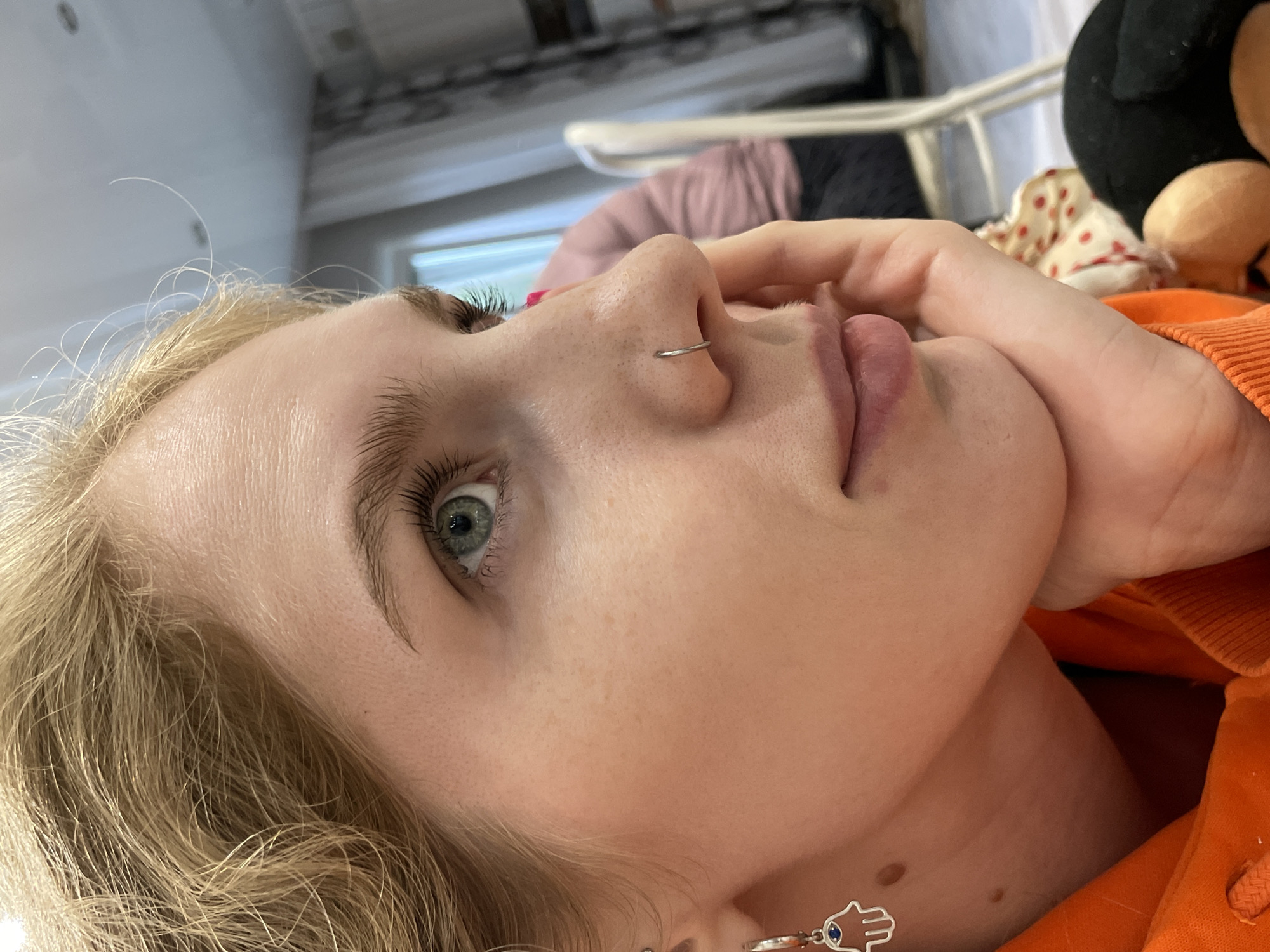 A close-up, slightly angled, indoor portrait captures a fair-skinned person with light blonde hair. A silver ring pierces their left nostril. Their eyes are a striking gray-green color. The person's face rests against their hand, giving the impression of contemplation. They wear an orange garment. An earring in the shape of a Hamsa dangles from their ear.
Behind the subject, out of focus, there are bedclothes and a stuffed animal. The setting is indeterminate; no information in the image suggests any specific location, let alone Izhevsk, Russia.