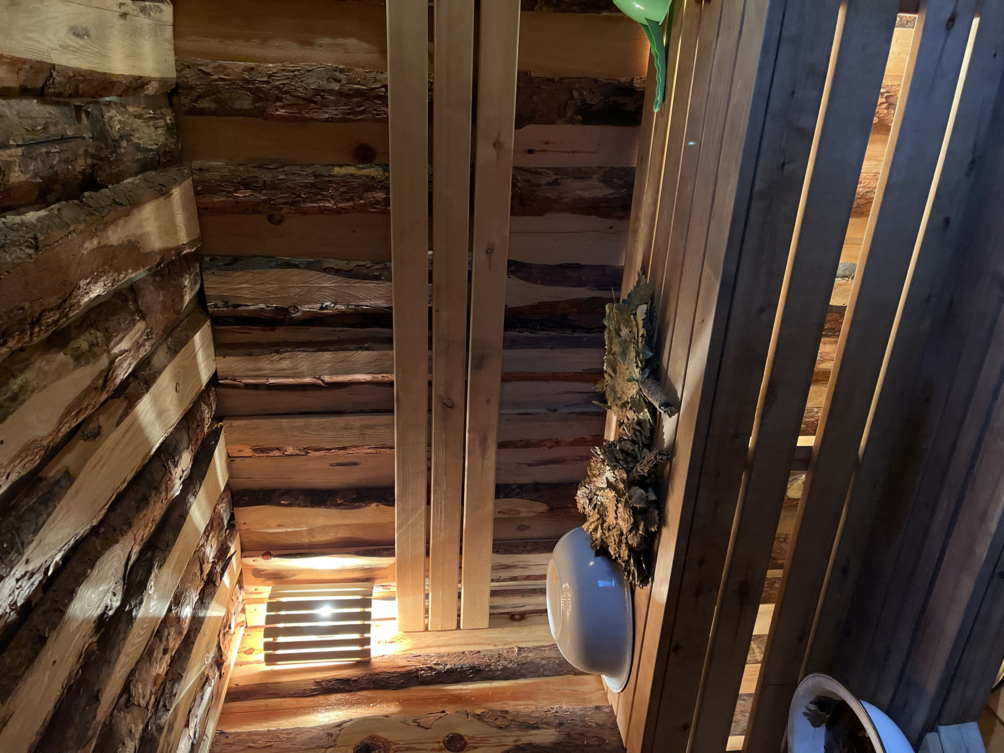 The image shows the interior of a wooden sauna or banya, possibly located in the city of Зыково, Russia. The walls are constructed from logs, and the seating or platforms are made of wooden planks. A wooden ventilation grate is seen in the wall. A birch whisk (вени́к) and a basin are mounted on the wall.