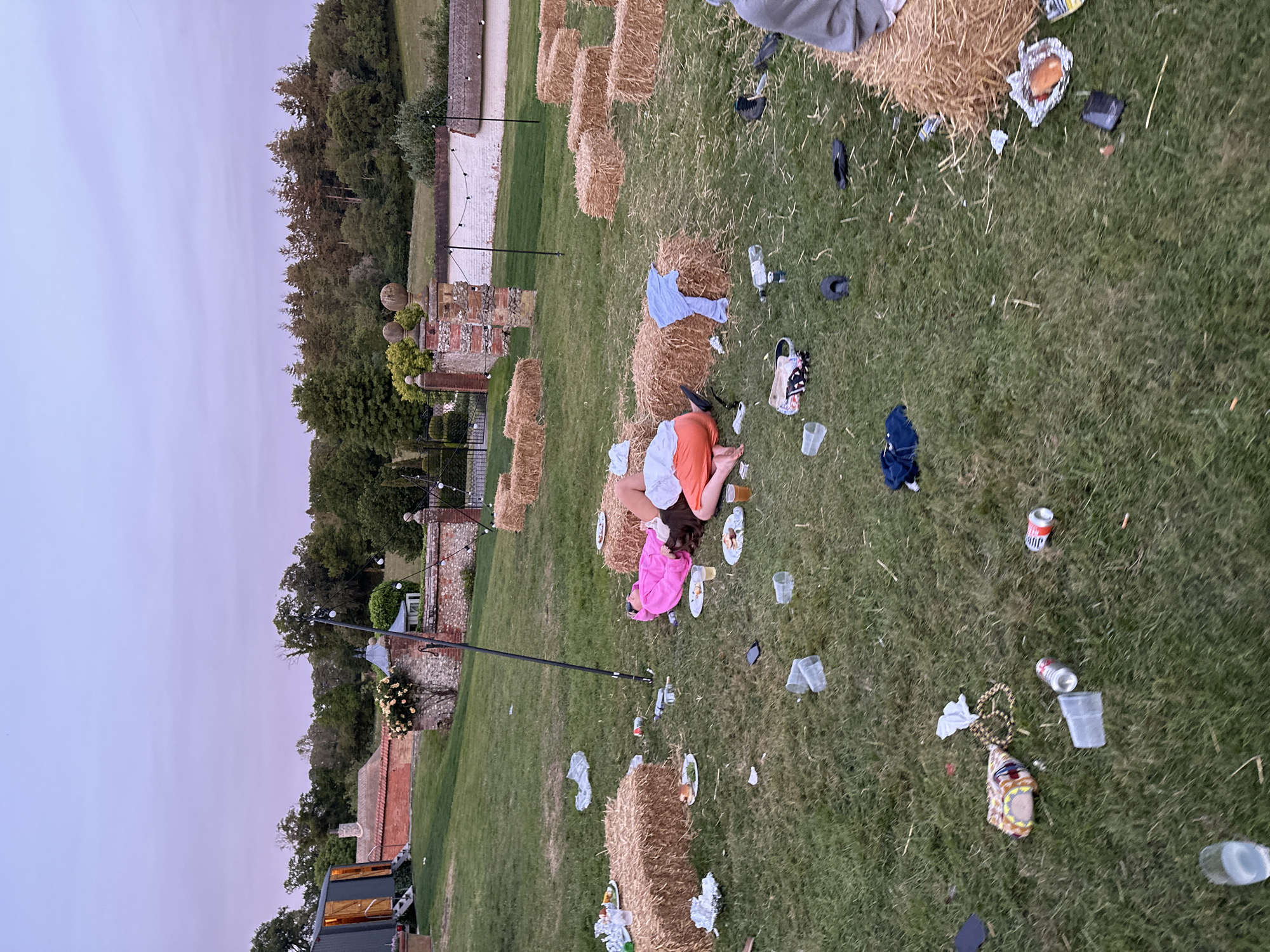 The image shows an outdoor event location littered with trash, including cans, cups, food wrappers, clothing, and personal items scattered across a grassy area. Bales of hay are positioned throughout the area, some bearing remnants of the event. Two individuals are lying on the ground, partially supported by the hay bales. In the background, a brick building and trees are visible under a dusk sky. The location is in the city of Unknown, Unknown.