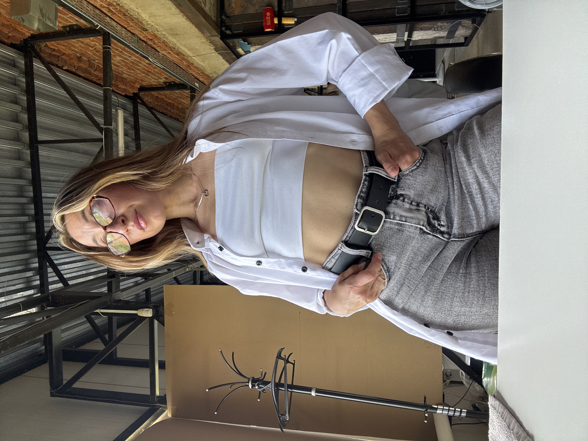 A woman with blonde hair and glasses, wearing a white tube top, an unbuttoned white shirt, and grey jeans with a black belt, stands in what appears to be a cafe or industrial space. She is looking to her left with her hands in her pockets. The background features exposed brickwork, metal beams, and a corrugated metal roof, suggesting an industrial aesthetic. A coat rack with a hanger is visible to the left of the woman. The image's lighting suggests it was taken during the day, possibly with natural light filtering in. It is not possible to determine the specific location within Chaikovsky, Russia, from the image alone.