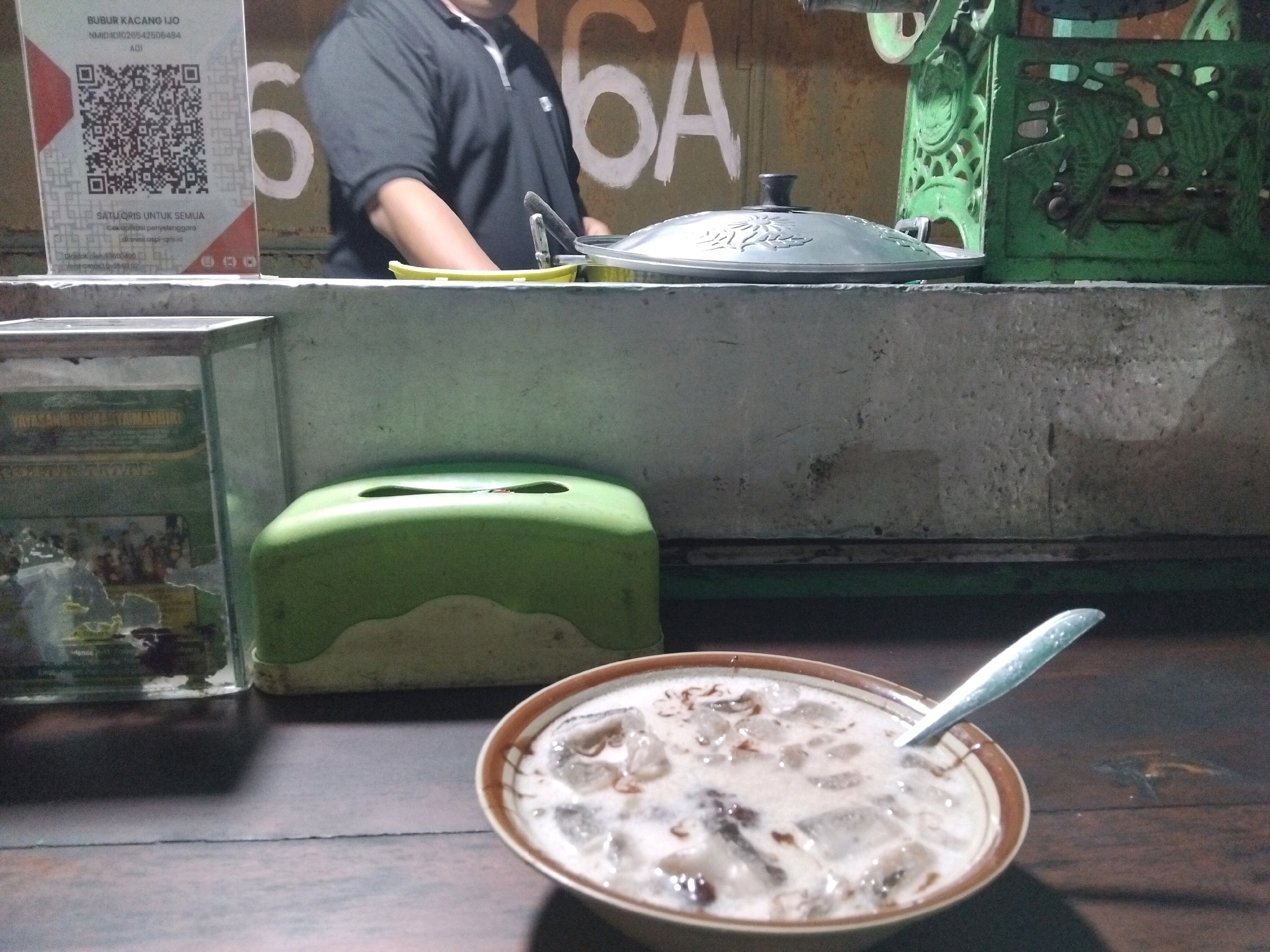An outdoor scene at what appears to be a food stall in Wedoro, Indonesia, shows a bowl of iced dessert with a spoon resting in it, placed on a wooden table. The dessert, likely bubur kacang ijo, is creamy white with visible ice cubes and darker bits, possibly beans or spices. To the left, a green tissue box sits on the table. In the background, a person in a black polo shirt is partially visible behind a counter displaying a QR code for payment. Behind the counter, metal containers and a decorative green metal structure are present. A wall with the painted numbers "6A" is visible in the upper background. The overall lighting suggests it is evening or dusk.