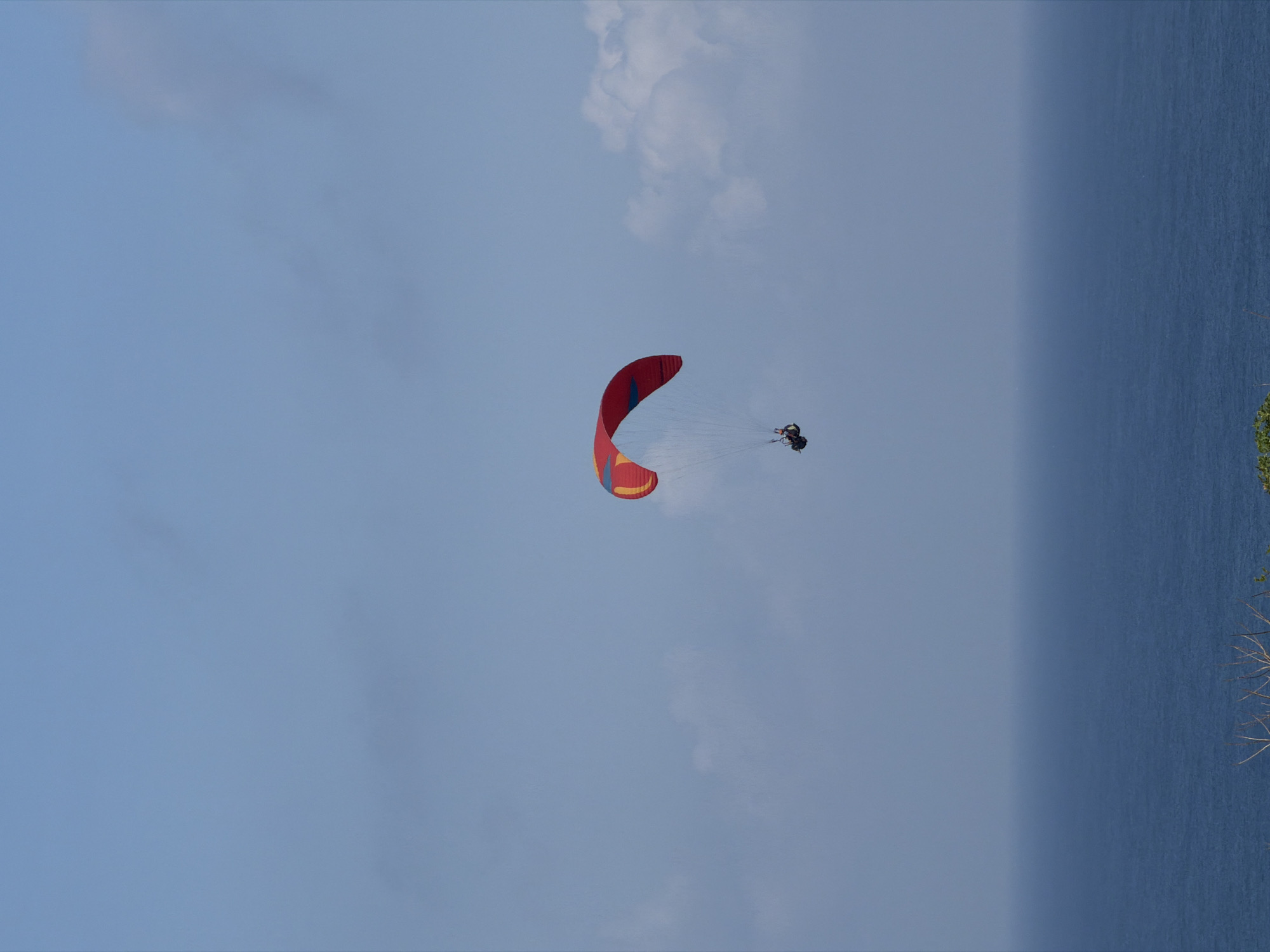 A paraglider with a red and yellow wing is soaring over the ocean. The paraglider is a small figure suspended beneath the wing, appearing to be a single person. The ocean is a deep blue and stretches to the horizon, where it meets a hazy sky with scattered white clouds. The scene is likely set near Kuta Selatan, Indonesia, a region known for its coastal landscapes and paragliding activities. The image captures a moment of freedom and adventure, with the vastness of the ocean and sky emphasizing the isolation and exhilaration of the activity.