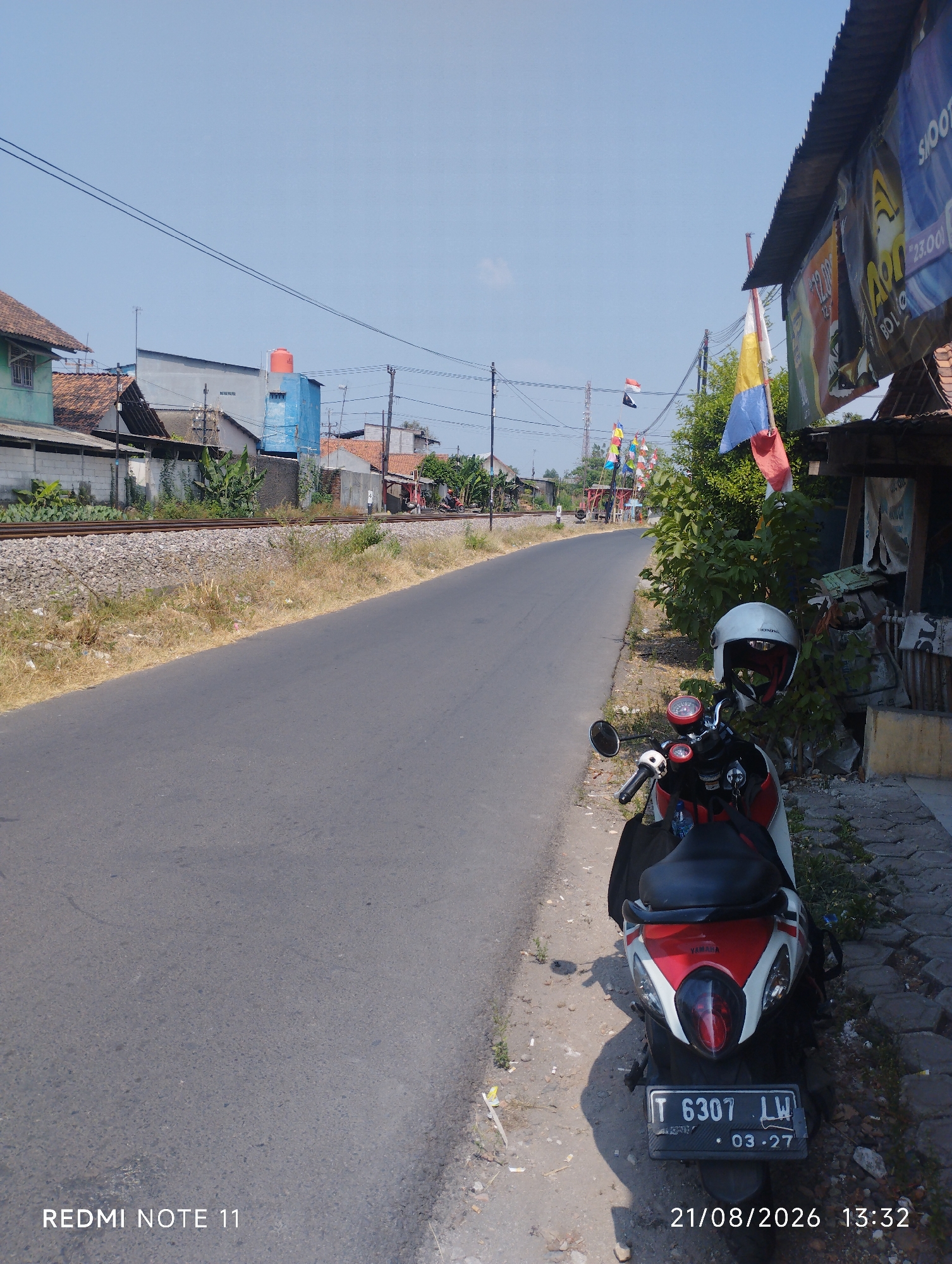 An outdoor, eye-level, full shot captures a street scene in Karawang, Indonesia. The asphalt road dominates the foreground and middle ground, stretching towards a distant bend under a clear blue sky. Along the left side of the road, a railway track runs parallel, bordered by dry grass and gravel. The background reveals residential buildings and structures, including a blue building with an orange water tank, and a few scattered trees.
To the right of the road, a motorcycle, predominantly white and red with a black seat, is parked on a paved sidewalk. A white helmet rests on the handlebars. The motorcycle's license plate reads "T 6307 LW" and "03-27". A corrugated metal roof overhang, adorned with colorful banners and flags, extends over a stall or shop area to the right. The scene appears to be captured during daylight hours, as indicated by the bright sunlight and the presence of shadows. The date and time "21/08/2026 13:32" are superimposed on the bottom right of the image, alongside the text "REDMI NOTE 11" on the bottom left.
