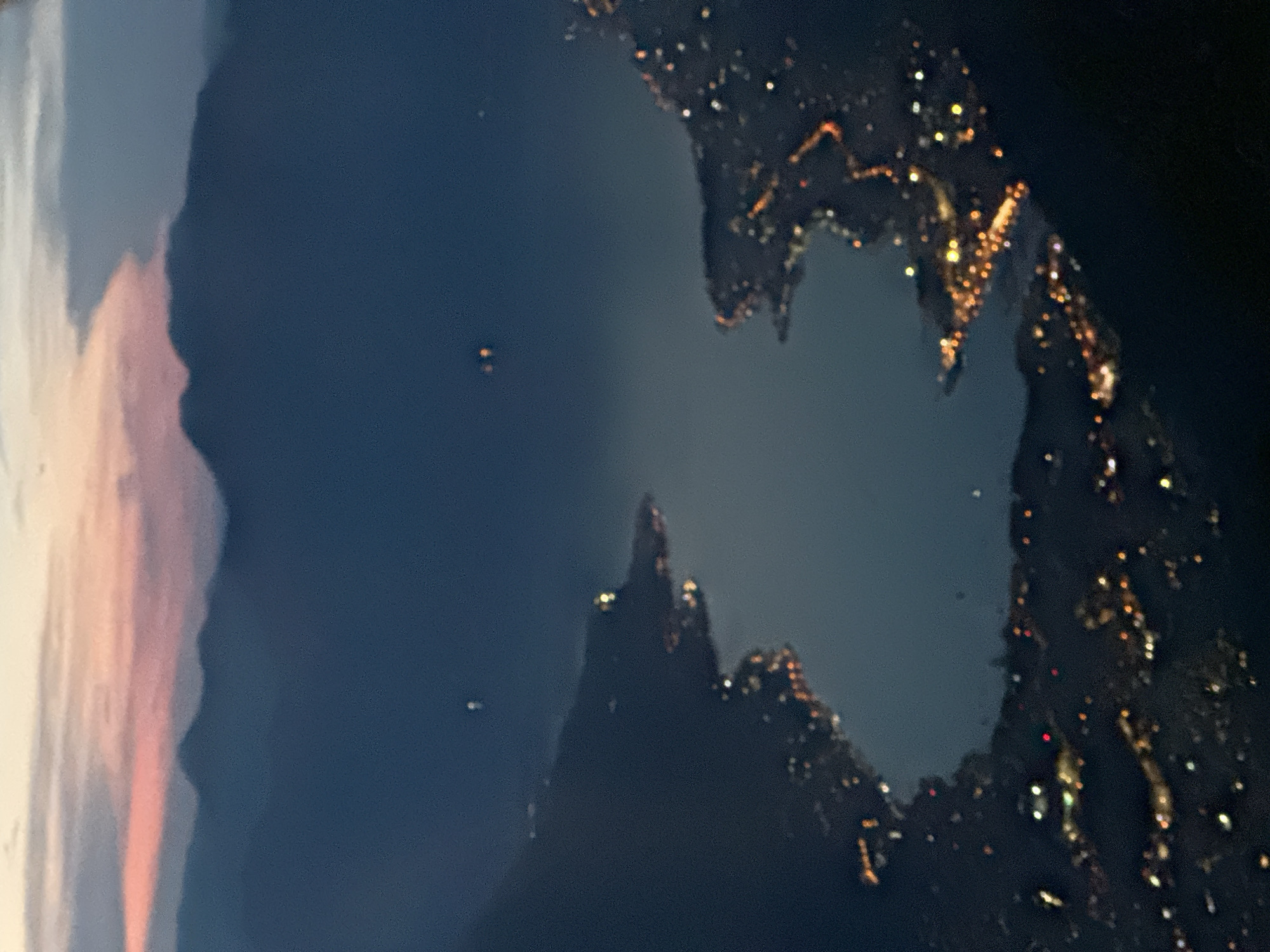 An aerial view captures the Bay of Kotor at dusk, with the lights of the coastal town of Dobrota, Montenegro, beginning to illuminate the landscape. The sky is painted with the soft hues of a fading sunset, with streaks of pink and orange clouds above a dark, silhouetted mountain range. The bay itself is a dark expanse of water, reflecting the distant lights and the twilight sky. Along the shore, clusters of warm, yellow and orange lights mark the developed areas of Dobrota, tracing the coastline. The buildings are not individually discernible due to the distance and low light conditions, but the presence of settlements is clear. The scene is tranquil, with the quietude of evening settling over the bay and its surroundings.