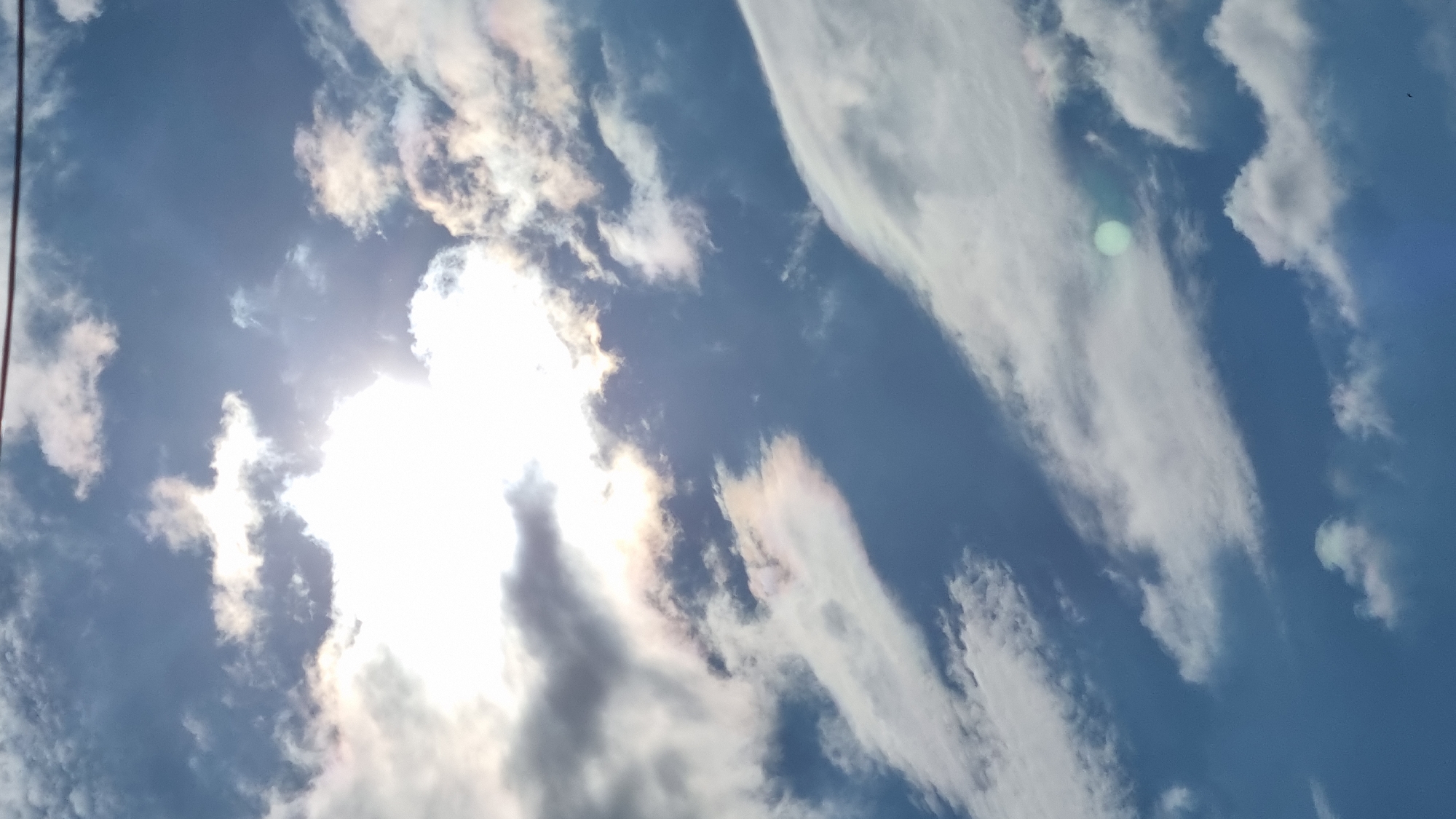 The image shows a sky with clouds and the sun. The sun is partially obscured by a large, bright cloud, creating a lens flare effect. There are smaller, wispy clouds scattered across the sky, some of which appear to have iridescent colors. A thin, dark line, possibly a power line, is visible at the top of the image. The scene is a typical sky view, with no specific landmarks or features that would indicate its location within Tbilisi, Georgia.