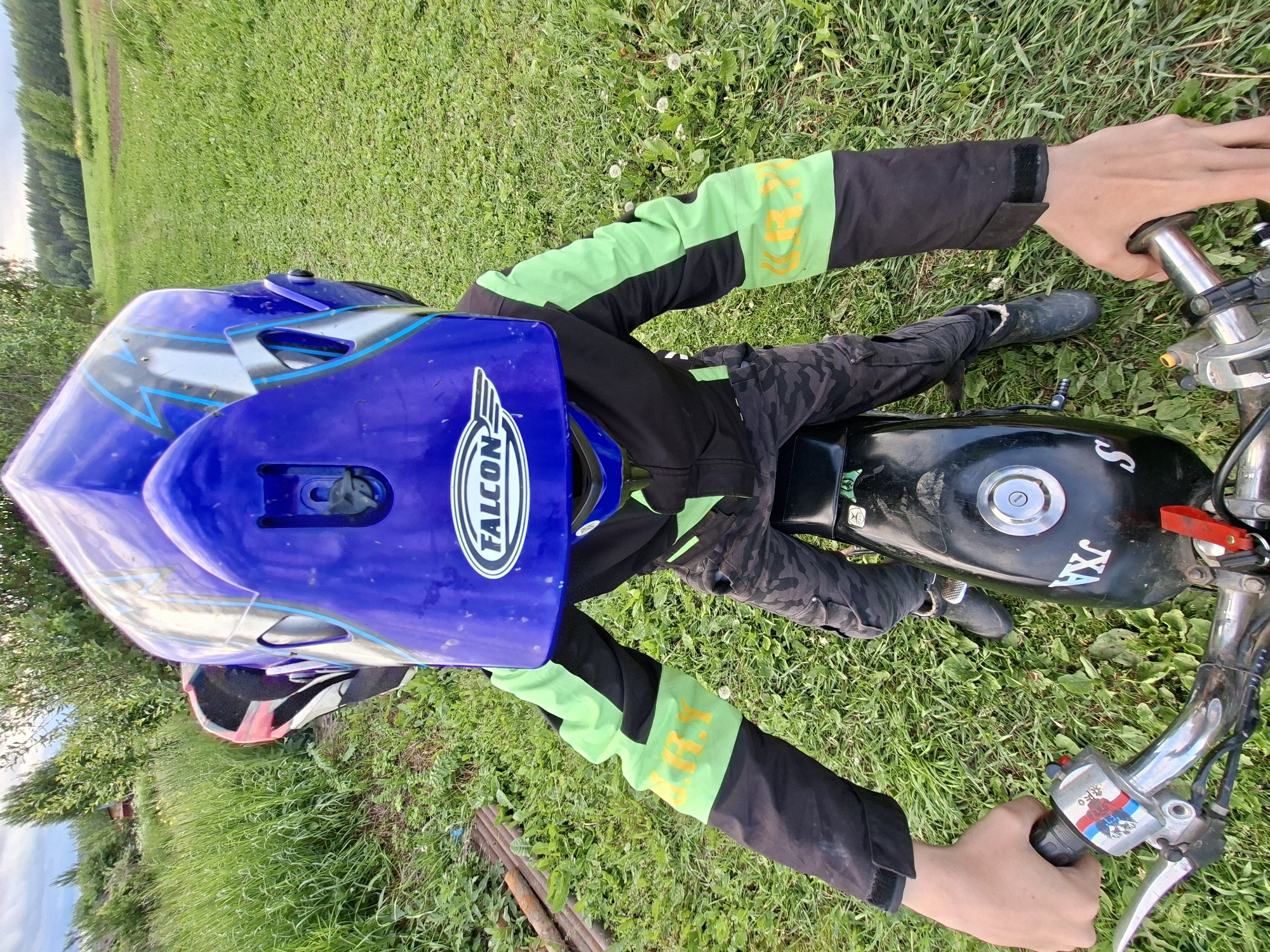 A first-person perspective image captures a person seated on a small, black motorcycle in an outdoor, grassy environment within the city of Unknown, Unknown. The individual is wearing a prominent blue full-face helmet adorned with silver and light blue lightning bolt graphics, featuring a white oval "FALCON" logo on the chin guard. They are dressed in a black and neon green long-sleeved riding jacket, with the right sleeve displaying yellow text, possibly "U.R.I.", on its green band. Dark camouflage cargo pants and dark boots complete the attire. Both light-skinned hands are gripping the motorcycle's chrome handlebars; the left hand is near a control cluster featuring a red button, a multi-colored (white, blue, red) striped sticker, and a silver brake lever, while the right hand grasps a black throttle grip.
The motorcycle has a black fuel tank with a silver cap and white lettering "JXA" visible on its side. The ground consists of uneven green grass mixed with various weeds and dandelions. To the left, taller grass and dense foliage are present, with a distant red structure partially visible. On the ground near the left side, several rusty metal pipes or rods are discarded. To the right, an expansive green field stretches towards a distant line of dark green forest. The sky above is overcast with grey clouds, indicating natural daylight conditions.