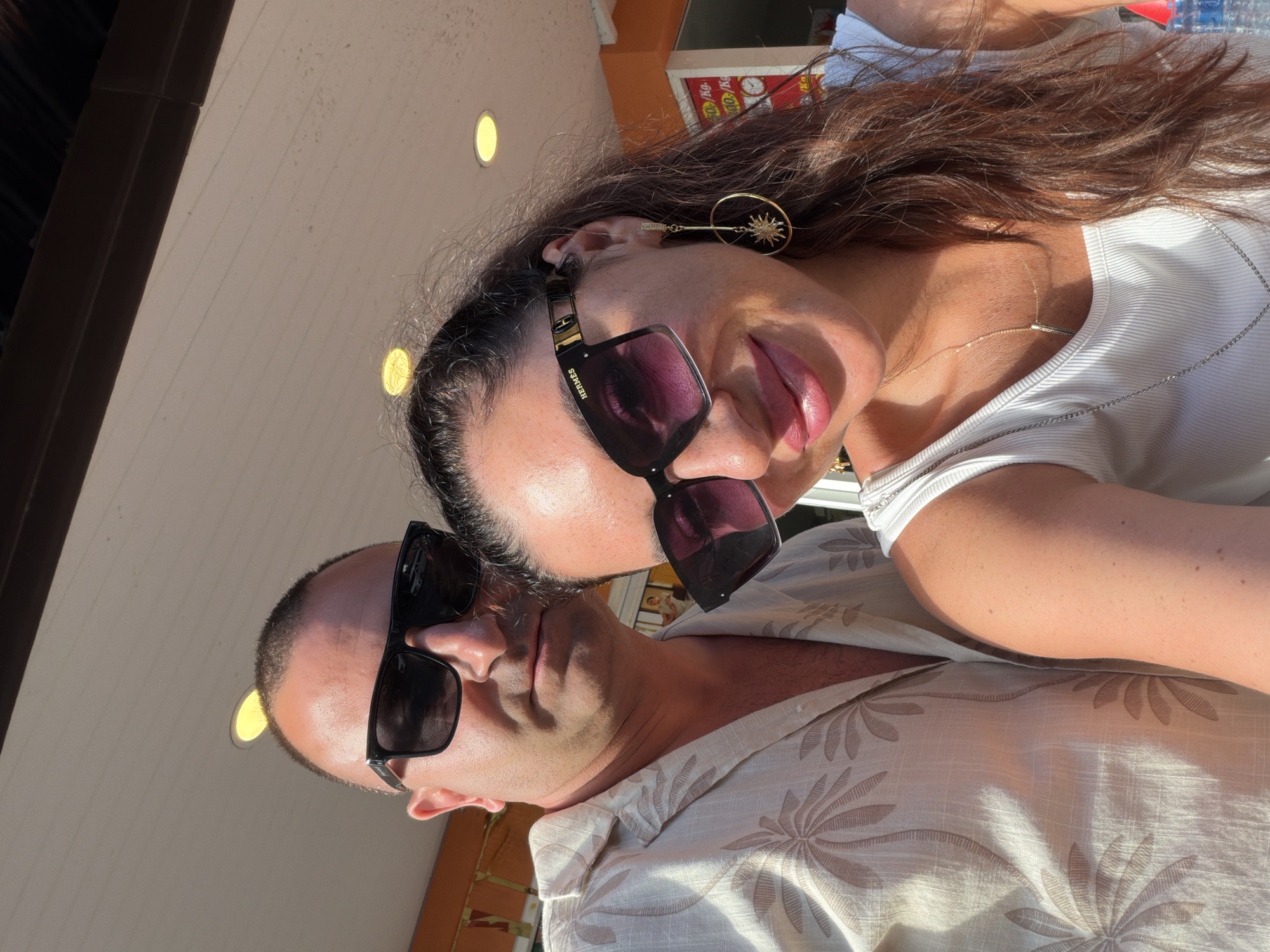 An overhead, close-up selfie shows a man and a woman in Patong, Thailand. The man, on the left, wears a tan, short-sleeved button-up shirt with a palm tree pattern. He has dark hair, is clean-shaven, and is wearing black sunglasses. The woman, on the right, leans into the frame, her head tilted and her eyes looking towards the camera. She has long, dark brown hair and is wearing a white tank top and large, black sunglasses. A gold hoop earring with a sun charm hangs from her right ear. Both individuals are smiling and appear to be outdoors, with a light-colored ceiling or awning visible overhead. The background suggests an outdoor establishment with orange accents and signs, potentially a shop or restaurant. The lighting indicates it is daytime, with sunlight casting shadows.