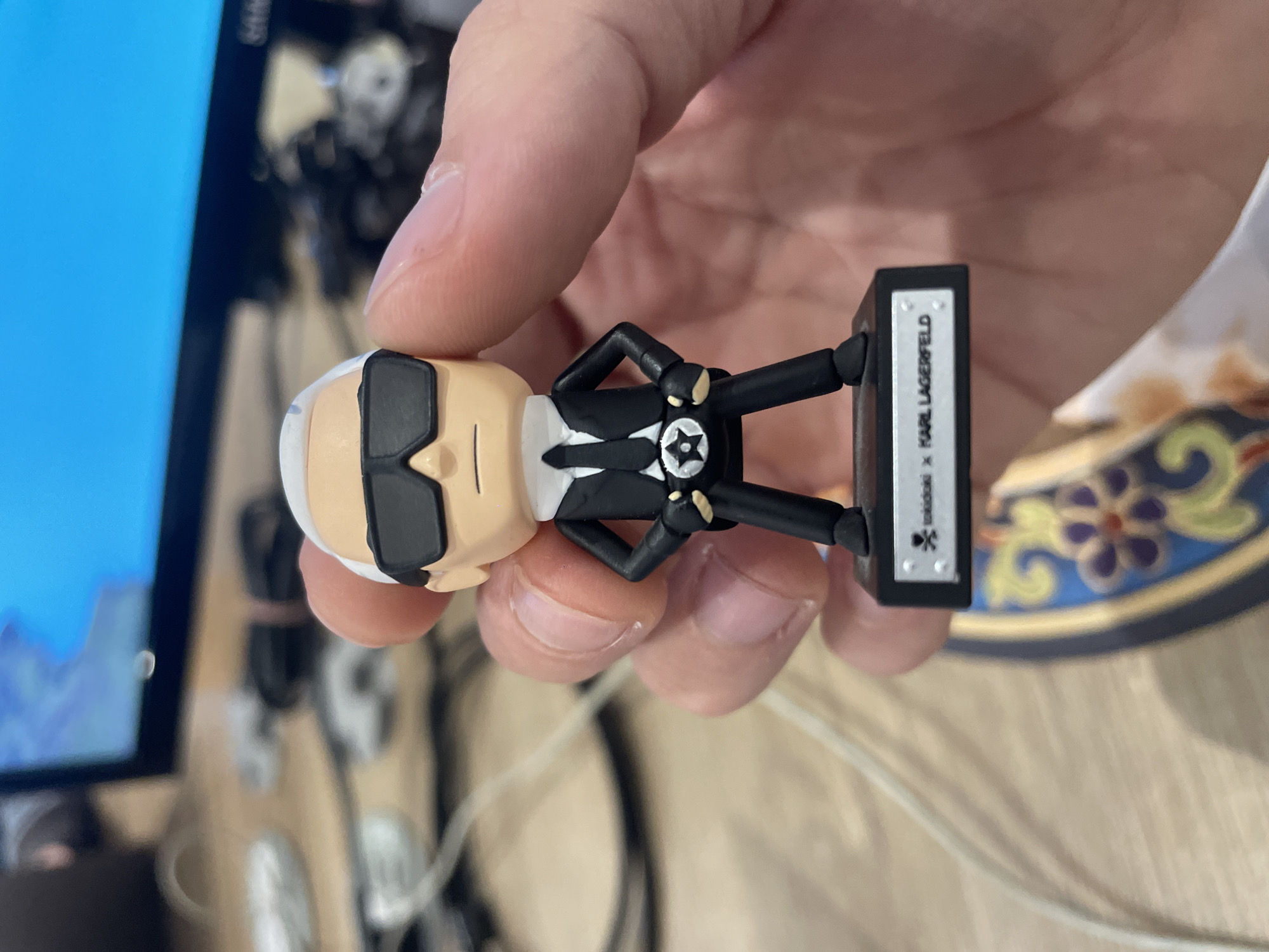 A hand holds a small Karl Lagerfeld figurine. The figurine is wearing a black suit, tie, and sunglasses. The base of the figurine is black and has text that appears to be the "Tokidoki x Karl Lagerfeld" collaboration logo. The background is slightly out of focus, but there is a computer monitor in the top left and a wooden table. The location is not discernible from the image.