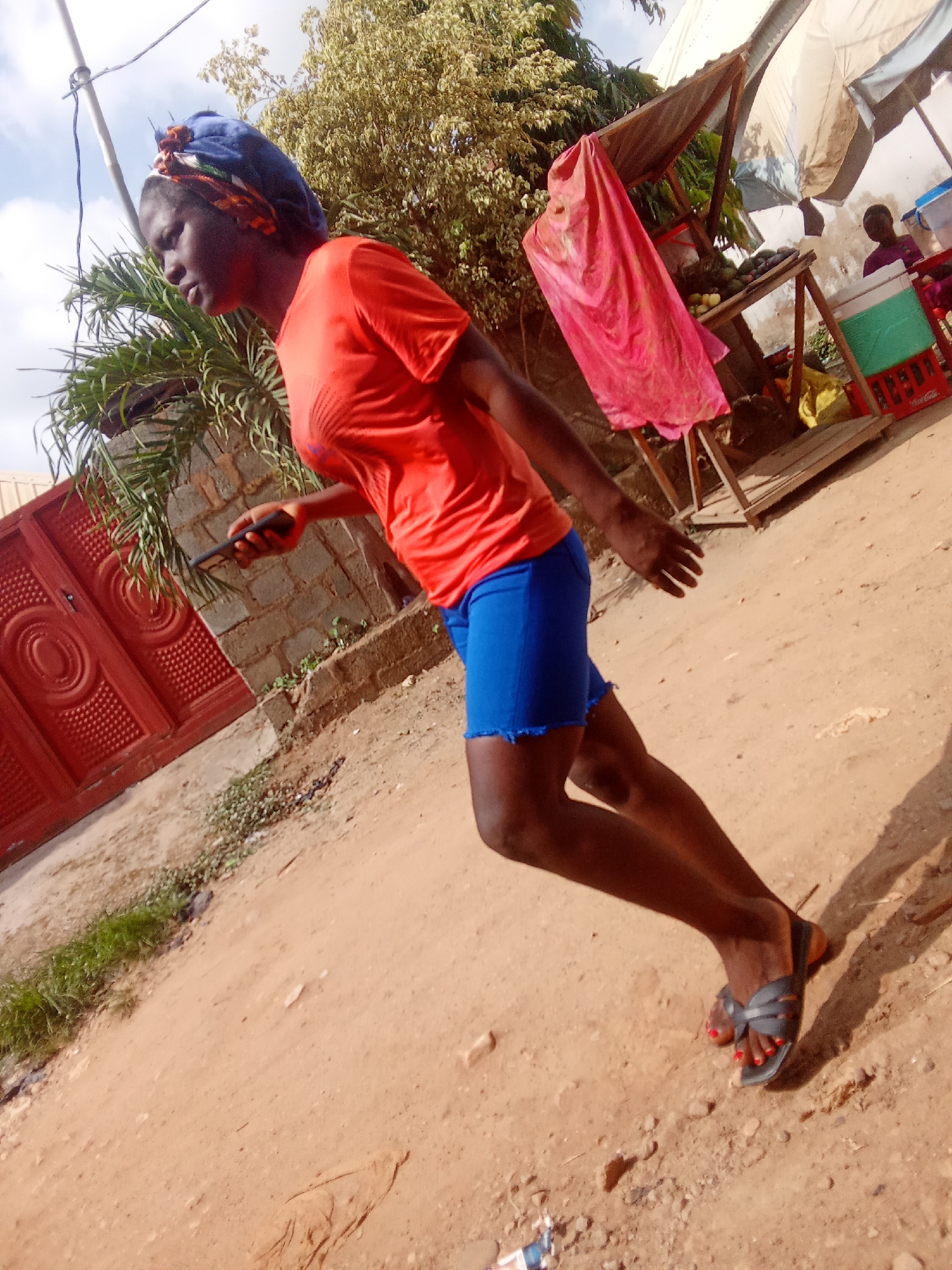 The image shows a young Black woman standing on a dirt path in Kagini, Nigeria. She is wearing an orange t-shirt, blue shorts, and dark sandals. Her hair is covered with a blue and patterned headscarf. She is holding a black smartphone in her right hand and looking downwards.
In the background, there is a market stall with a canopy, displaying some fruits and vegetables. A pink cloth is draped over one of the stalls. To the left of the woman is a red, ornate metal gate. Further in the background, another person is visible at the market stall. The scene suggests a bright, sunny day, possibly late morning or early afternoon, given the strong shadows. The overall setting is a vibrant, outdoor marketplace in a Nigerian community.