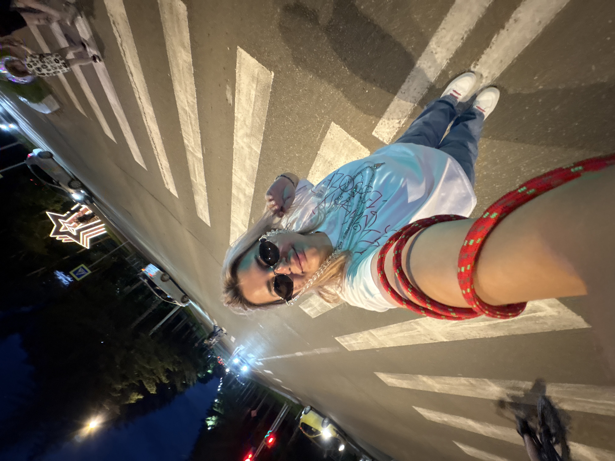 The image shows a woman taking a selfie at night on a crosswalk in Birobidzhan, Russia. The woman is wearing sunglasses and a white t-shirt. She is holding a red rope wrapped around her arm. In the background, there are cars and streetlights. There is also a decorative star illuminated with lights. The crosswalk is marked with white stripes on the asphalt. The sky is dark blue, indicating it is nighttime.
