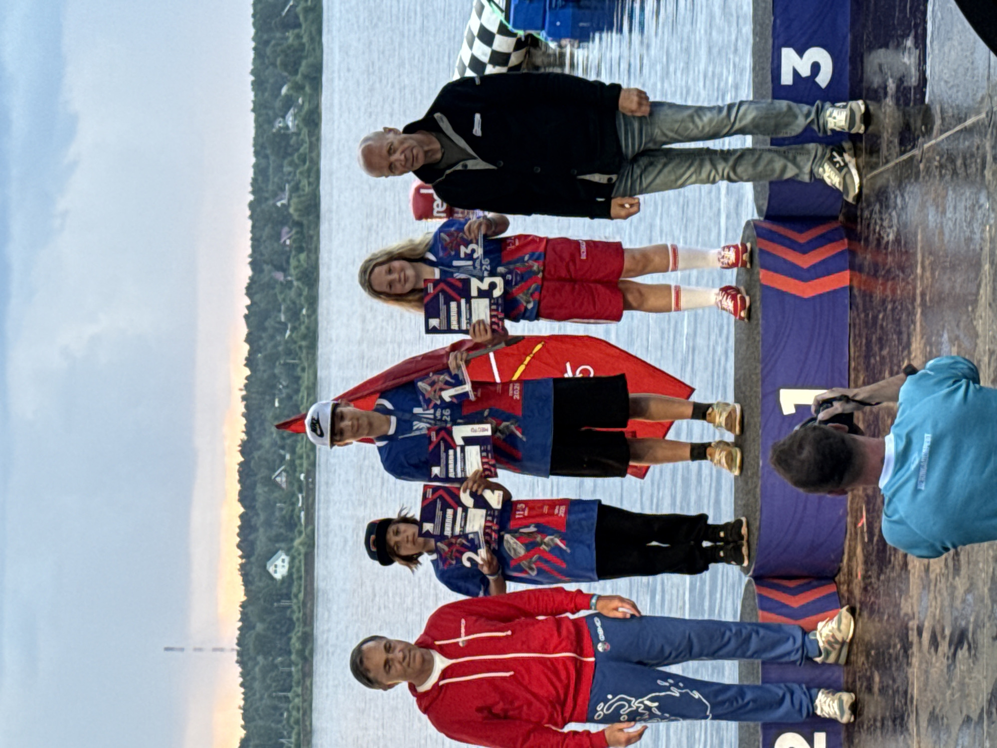 In Perm, Russia, at a waterside event, five individuals stand on a blue and purple podium. On the far left, a man in a red hoodie and blue pants with white designs is looking towards the camera. Next to him, a young person wearing a blue shirt, black pants, and a black baseball cap holds two awards marked with the number "2." To their right, a young male in a blue shirt and black shorts, wearing a white Nike baseball cap, holds an award marked with "1." Next to him, a young female in a blue and red shirt and red shorts, with blonde hair, holds an award marked with "3." On the far right, a man in a black jacket and blue jeans stands with a neutral expression. Behind them, a body of water stretches towards a treelined horizon under a cloudy sky. A checkered inflatable object is visible in the water to the right of the podium. In the foreground, a person in a turquoise shirt is taking a photograph with a camera. The podium has numbers "1," "2," and "3" displayed. The event appears to be an awards ceremony for a water sport.