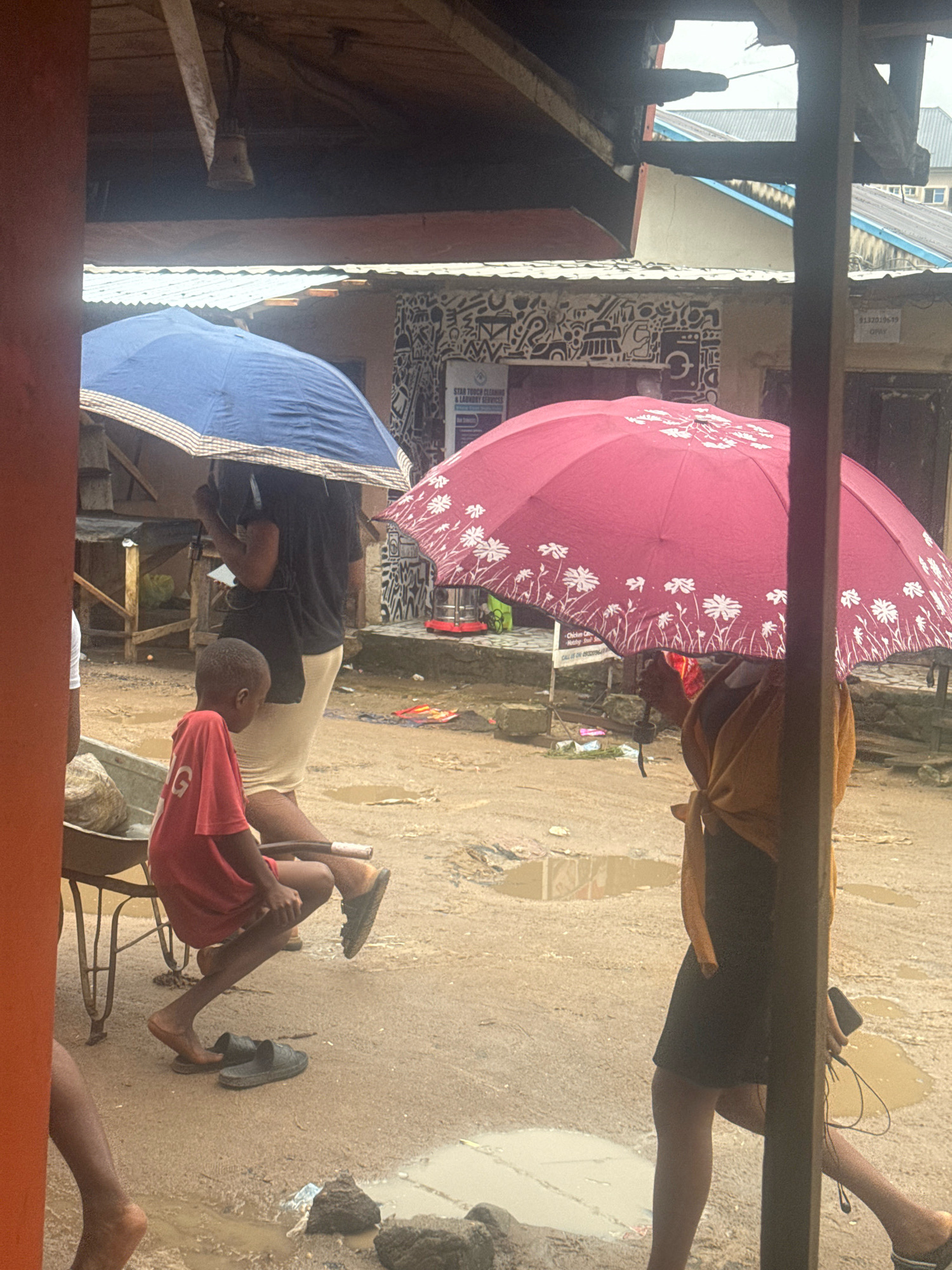 The image depicts a rainy day in what appears to be a marketplace or outdoor area in Ihiagwa, Nigeria. In the foreground, a young boy wearing a red t-shirt is sitting on a wheelbarrow, looking to his right. His legs are bare, and a pair of dark sandals are placed on the ground near him. To his left, another person's bare legs are visible.
Behind the boy, a person is holding a blue umbrella, obscuring their face and much of their body. They are wearing a dark shirt and a light-colored skirt or pants. To the right, another individual is holding a large, pink umbrella decorated with white floral patterns. This person is wearing a dark dress and an orange wrap around their shoulders, and they are holding a mobile phone with wired earphones.
The ground is wet and muddy, with several puddles scattered throughout the scene, indicating recent or ongoing rainfall. In the background, there are structures that suggest shops or stalls, with corrugated metal roofs and some visible signage, one with the text "STAR TOUCH CLEANING & LAUNDRY SERVICES". The overall atmosphere is one of a typical day in a Nigerian city during the rainy season, with people using umbrellas for protection from the elements.