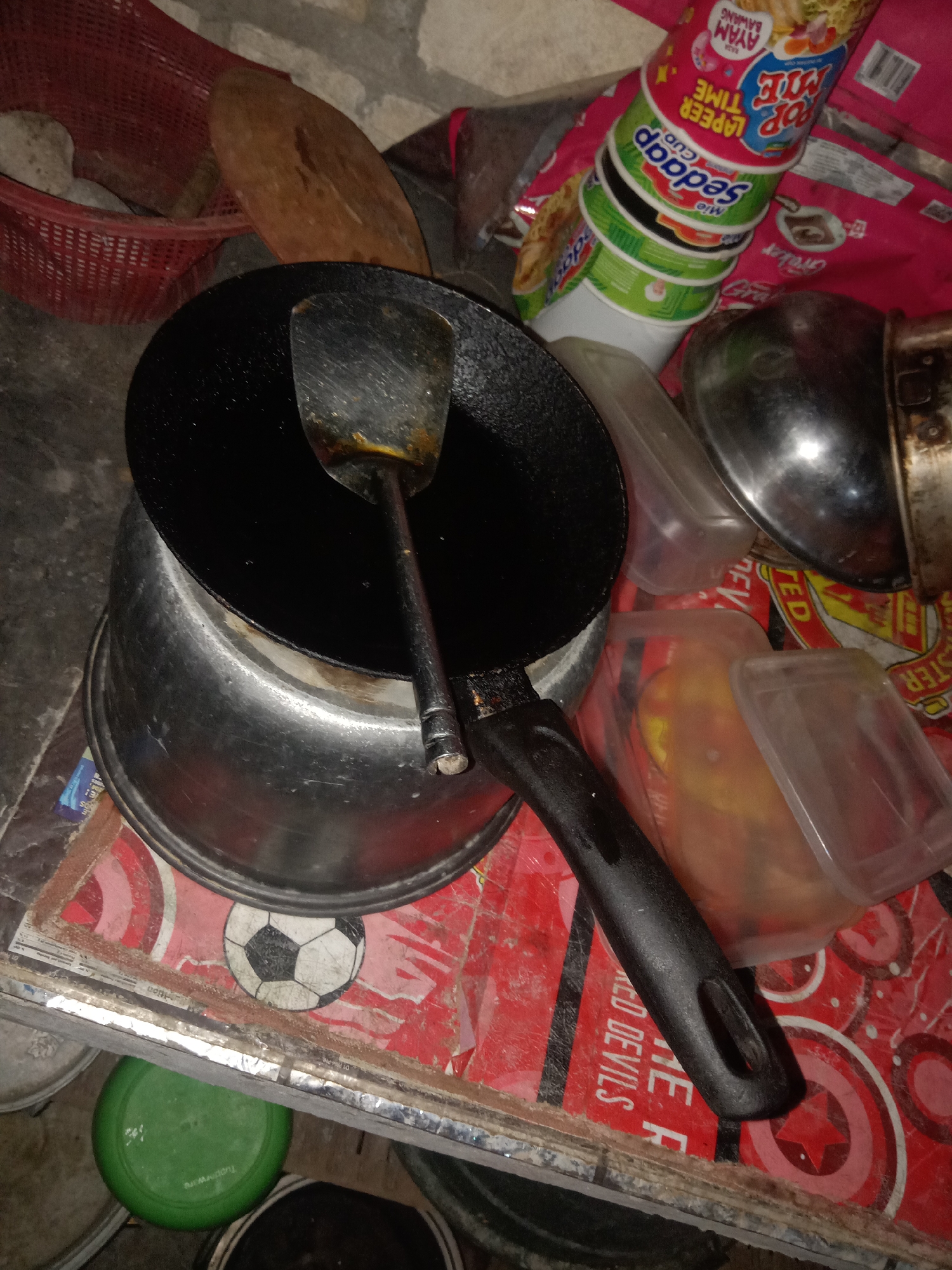 The image depicts a cluttered kitchen scene, likely in Jumput, Indonesia. In the foreground, a black frying pan sits atop a metal pot. A metal spatula is partially submerged in the pan. To the right, several containers are stacked, including a clear plastic container holding some food item and a stack of instant noodle cups. Behind these, a silver bowl is visible. The entire arrangement is placed on a surface covered with a red and white patterned material, featuring a soccer ball graphic. To the left, a red basket contains some items, and a round wooden cutting board is also present. Various other objects and containers are scattered around the scene, suggesting a working or storage area. The lighting is dim, creating shadows and highlighting the textures of the objects.