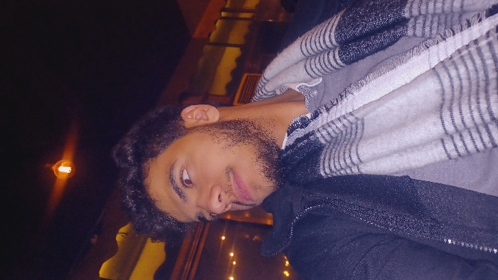 A person with curly hair and a beard faces the viewer. They wear a striped scarf over a dark jacket. The background is dark, with warm, yellow lighting in the lower portion and a blurred lightbulb visible on the left. A structural element, possibly a window frame, is visible behind the person. The image is rotated counter-clockwise.
