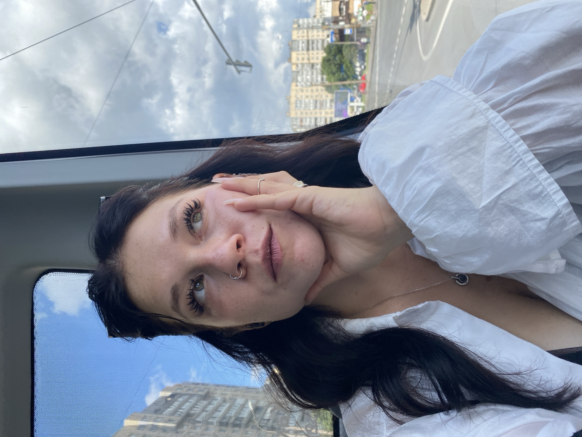 A young woman with dark hair and long eyelashes is sitting in a vehicle, looking out the window. She is wearing a white button-down shirt and a necklace. Her left hand is resting on her cheek. Outside the window, a cityscape is visible with buildings, a road, and a cloudy sky. The scene appears to be in Saint Petersburg, Russia, as indicated by the architecture and the presence of streetlights characteristic of the city.