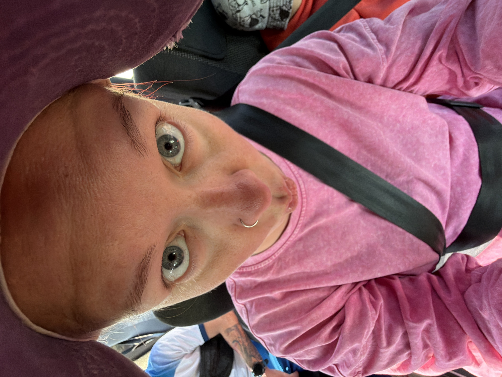 A selfie taken from an upside-down perspective, likely within a vehicle. A person with fair skin, greyish-blue eyes, and a nose ring is prominently featured. They are wearing a pink, textured long-sleeved shirt and a black seatbelt. Parts of two other people can be seen in the background. The setting appears to be indoors, possibly in a car. There is no contextual information to identify the location as Chaikovsky, Russia.