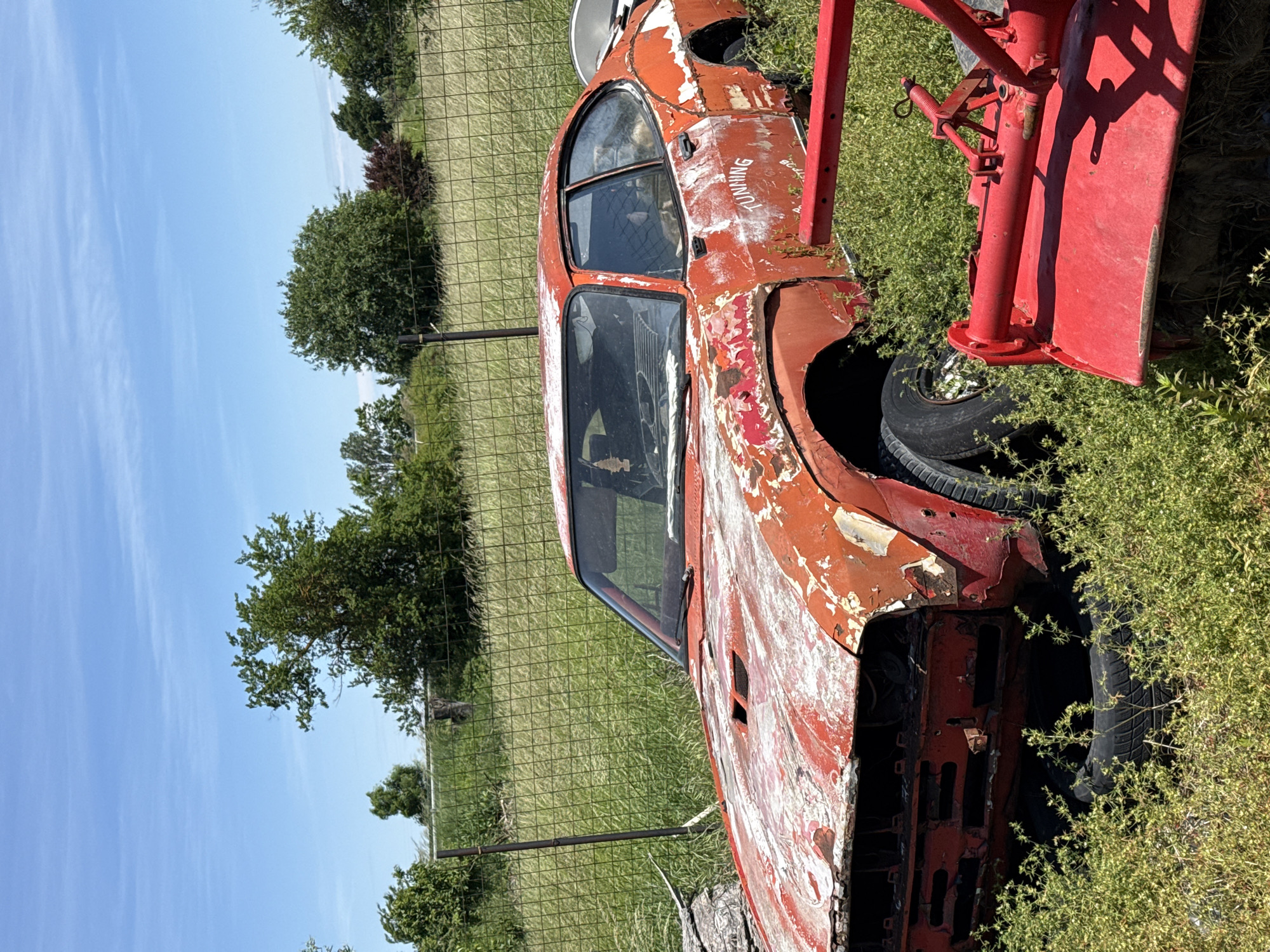 An abandoned, rusted-out red car, with "TUNING" spray-painted on the door, is surrounded by grass and positioned near a wire fence. The car's front is missing, revealing stacked tires. A red piece of heavy machinery is located nearby. The sky is blue. The location is Omurtag, Bulgaria.