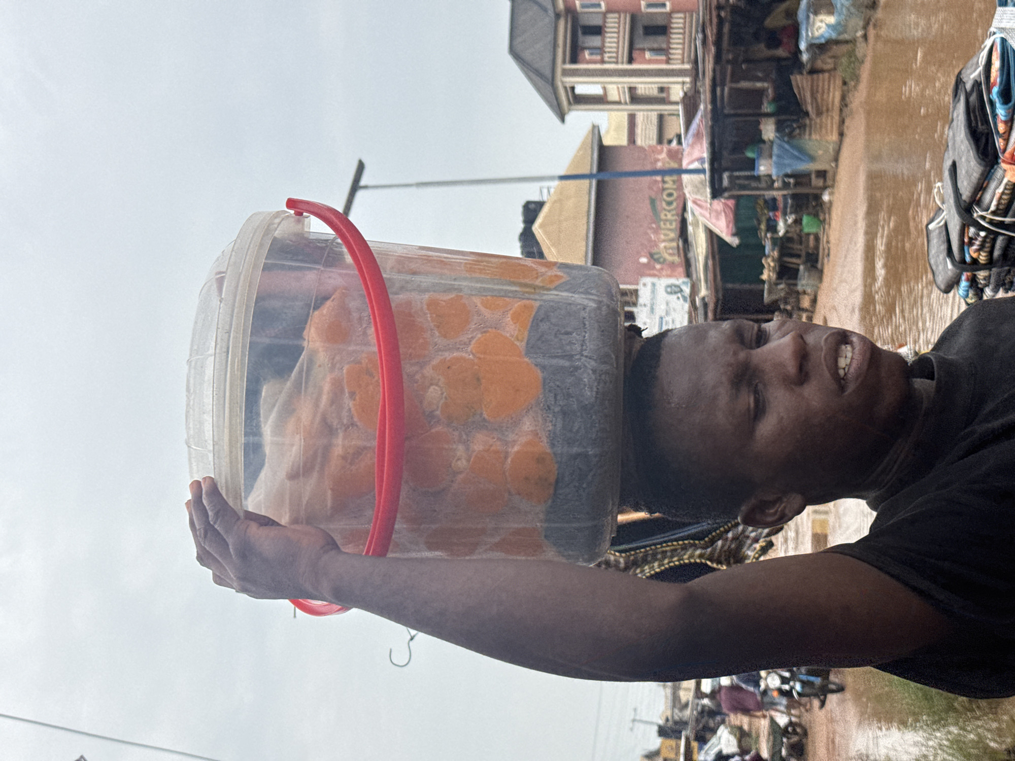 A young Black male is depicted in a medium close-up shot, carrying a clear plastic bucket filled with orange and grey materials on his head. He is wearing a black t-shirt and has his right arm raised, supporting the bucket. The background suggests an outdoor market setting in Ihiagwa, Nigeria, with stalls, various goods, and people visible. The ground is wet and muddy, indicating recent rainfall. The sky is overcast, contributing to the overall muted lighting of the scene. The man's facial expression is neutral, and his mouth is slightly open. In the foreground, a stack of folded fabric or mats is partially visible. The overall impression is one of daily commerce in a bustling Nigerian marketplace.