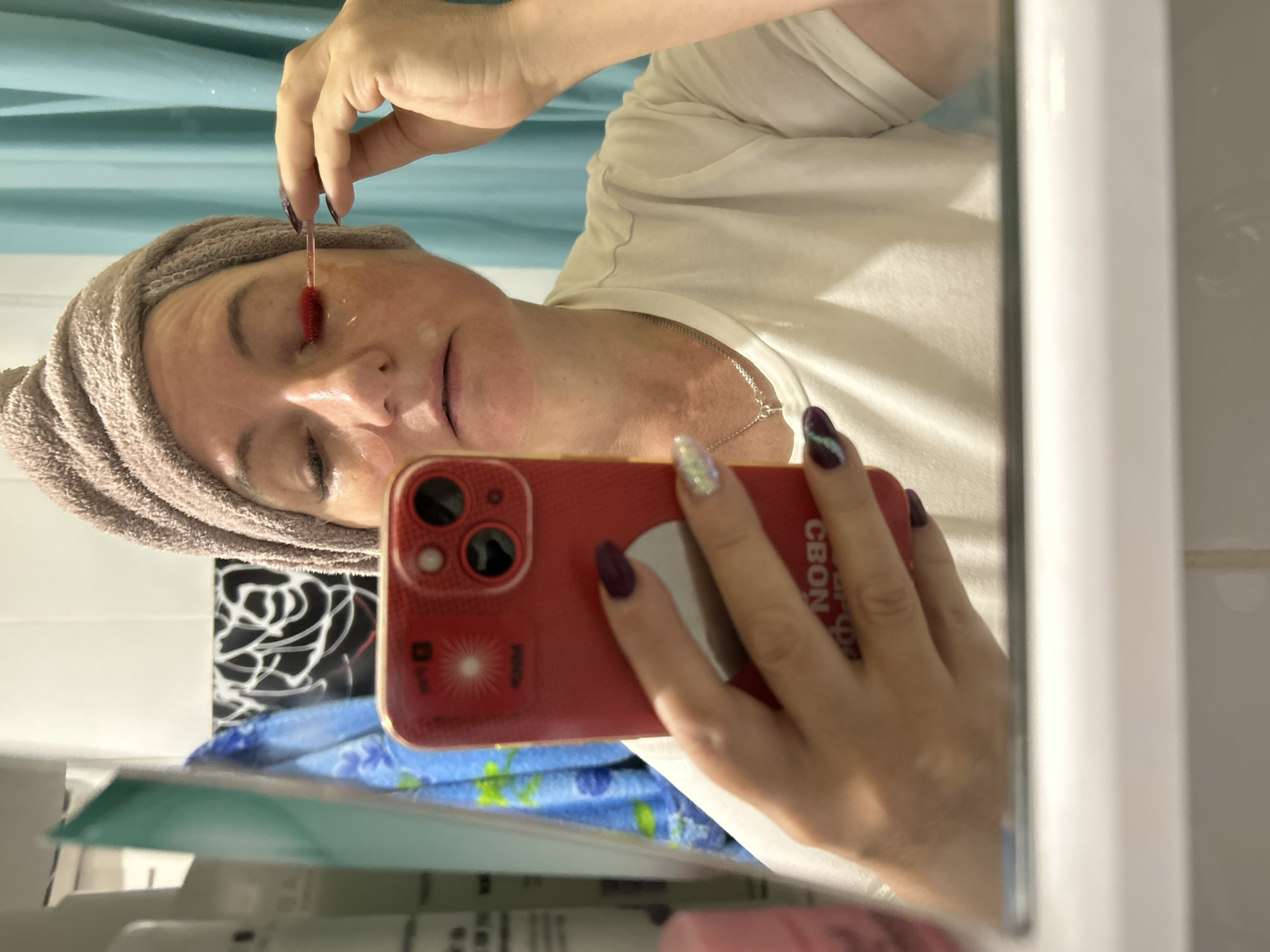 A woman, with a towel wrapped around her head, is using a red lash comb to apply product to her lashes while holding a red iPhone in her left hand, taking a selfie in a bathroom mirror. She is wearing a white t-shirt and a silver necklace. The bathroom appears to have teal blue curtains and a tiled wall. The image is a reflection in a mirror.