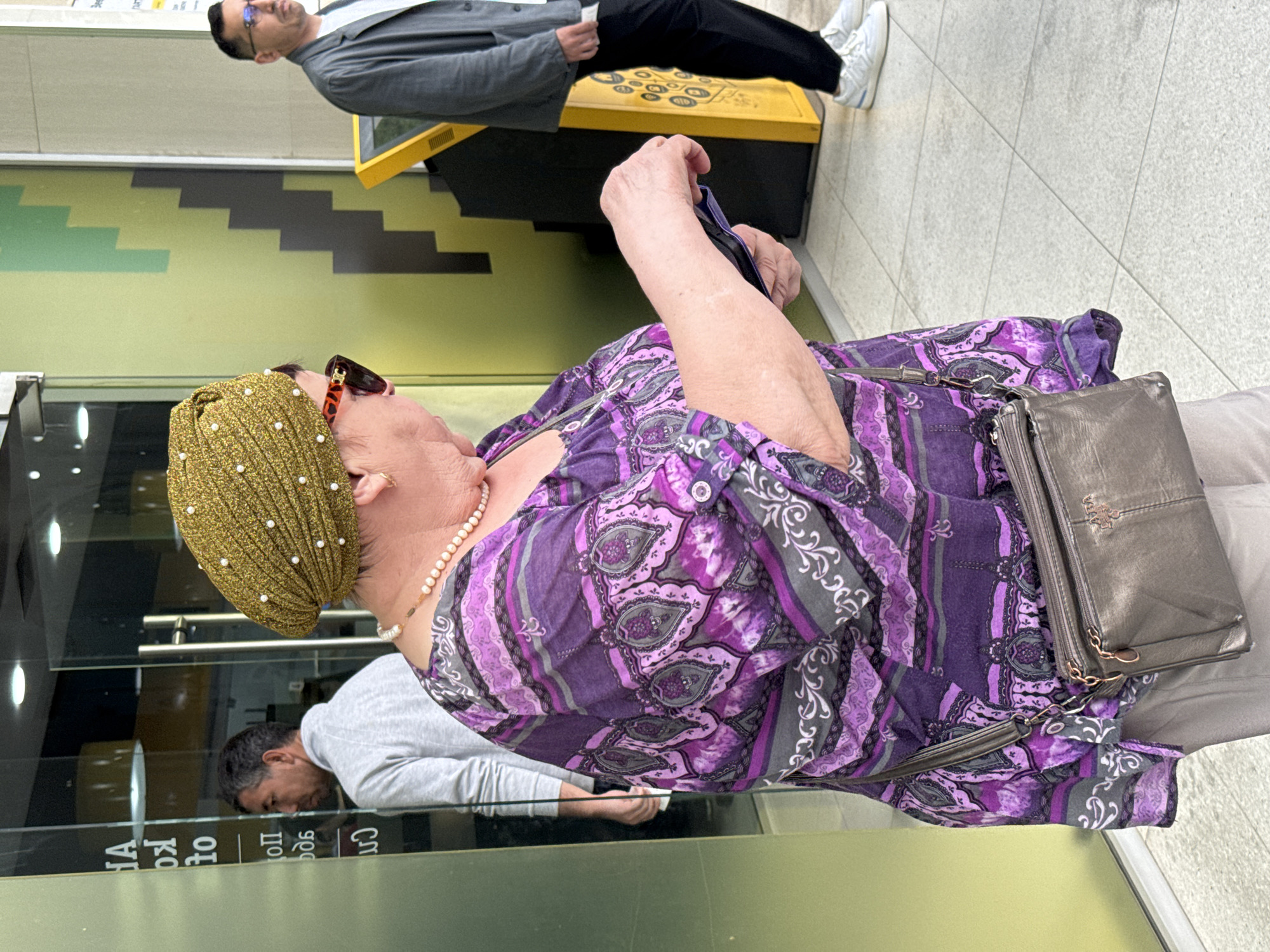 An eye-level, medium shot depicts a woman in a purple paisley blouse and a gold, beaded turban, with a grey purse over her shoulder, standing in a lobby in what is most likely Tashkent, Uzbekistan. Her reflection and that of a man in a grey long-sleeve shirt are visible in a glass pane in front of her. Behind her and in the upper left of the shot is an interior wall painted yellow and green. A man wearing glasses, a grey jacket, and black pants is standing behind what appears to be a payment kiosk with a yellow surface, in the upper part of the shot. The floor is tiled in light grey.
