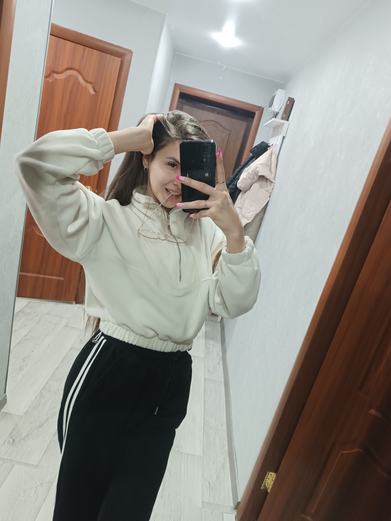 An indoor mirror selfie depicts a young adult female in what appears to be a residential hallway or entryway in Unknown, Unknown. The female subject, with long, dark brown hair, is wearing an off-white long-sleeved quarter-zip pullover top and black athletic pants with two white vertical stripes on the outer seam of the visible left leg. Her fingernails are painted bright pink, and she wears a small hoop earring on her visible left ear, along with two rings on her left hand. A pair of wire-frame eyeglasses is suspended from the zipper pull of her pullover. She is holding a black smartphone with her left hand to take the photograph, while her right hand is raised, with fingers partially within her hair. The subject is smiling. The background, reflected in the mirror, shows light-colored walls, light wood-look flooring, and two dark brown wooden doors with panel designs. To the right, a white wall shelf holds items, and below it, multiple garments, including a light beige puffer jacket, hang on hooks. A ceiling light fixture is reflected in the upper portion of the mirror.