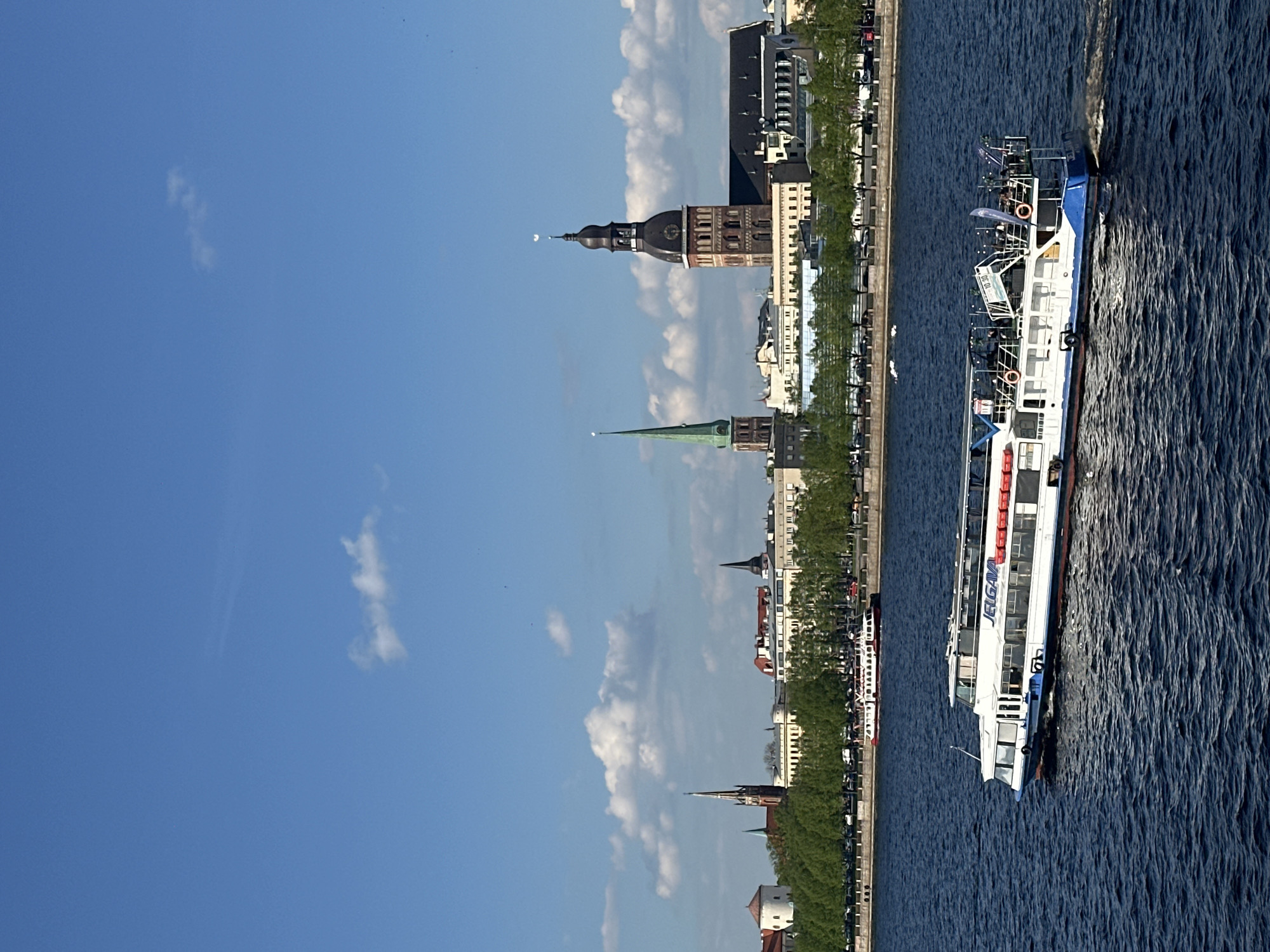 The image depicts a view of the skyline of Riga, Latvia, from the Daugava River. Several iconic buildings are visible, including the spires of St. Peter's Church and Riga Cathedral, characteristic of the city's architecture. Along the riverbank, there are trees and buildings. A tour boat named "Jelgava" is seen on the river. The sky is blue with some white clouds, suggesting daytime.