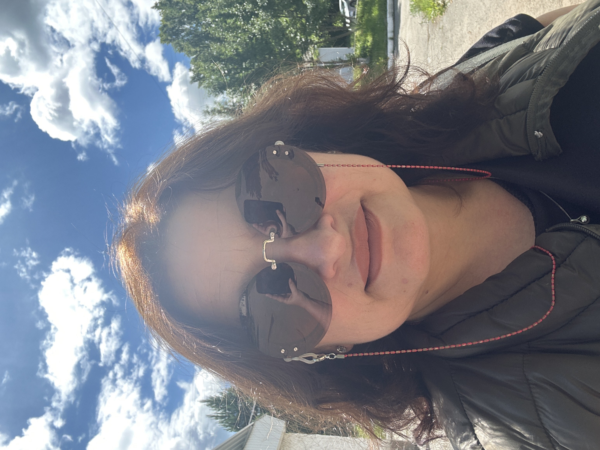 A medium shot photograph captures a woman wearing sunglasses and a dark vest. She is standing outdoors on a sunny day, with a bright blue sky and white clouds visible in the background. The woman has dark, wavy hair and is looking towards the camera. Her sunglasses have a reflective lens, showing a partial reflection of someone holding a phone to take the picture. A thin, beaded string, likely for glasses, is attached to the side of her sunglasses and drapes down her chest. The woman is wearing a dark vest over a dark top. The environment appears to be an urban or suburban setting, with hints of trees and a building in the background, and a paved surface in the foreground. The specific location is the city of Uchaly, Russia.