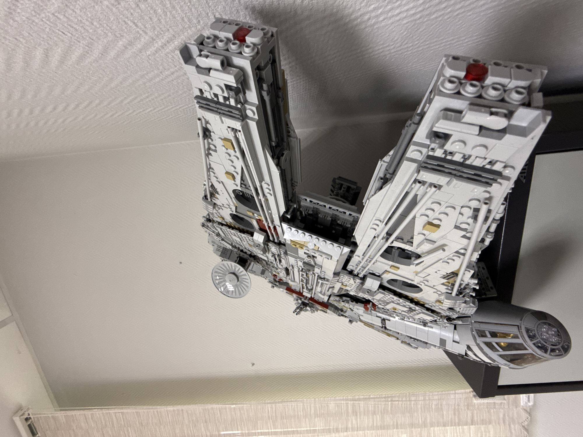 The image shows a Lego model of the Millennium Falcon, a fictional spacecraft from the Star Wars franchise. The model is predominantly light gray and white, with some red and black details. It is mounted on a black stand. The background includes a white textured ceiling and a glimpse of a curtain or window covering. There is no indication the image was taken in Krasnoyarsk, Russia.
