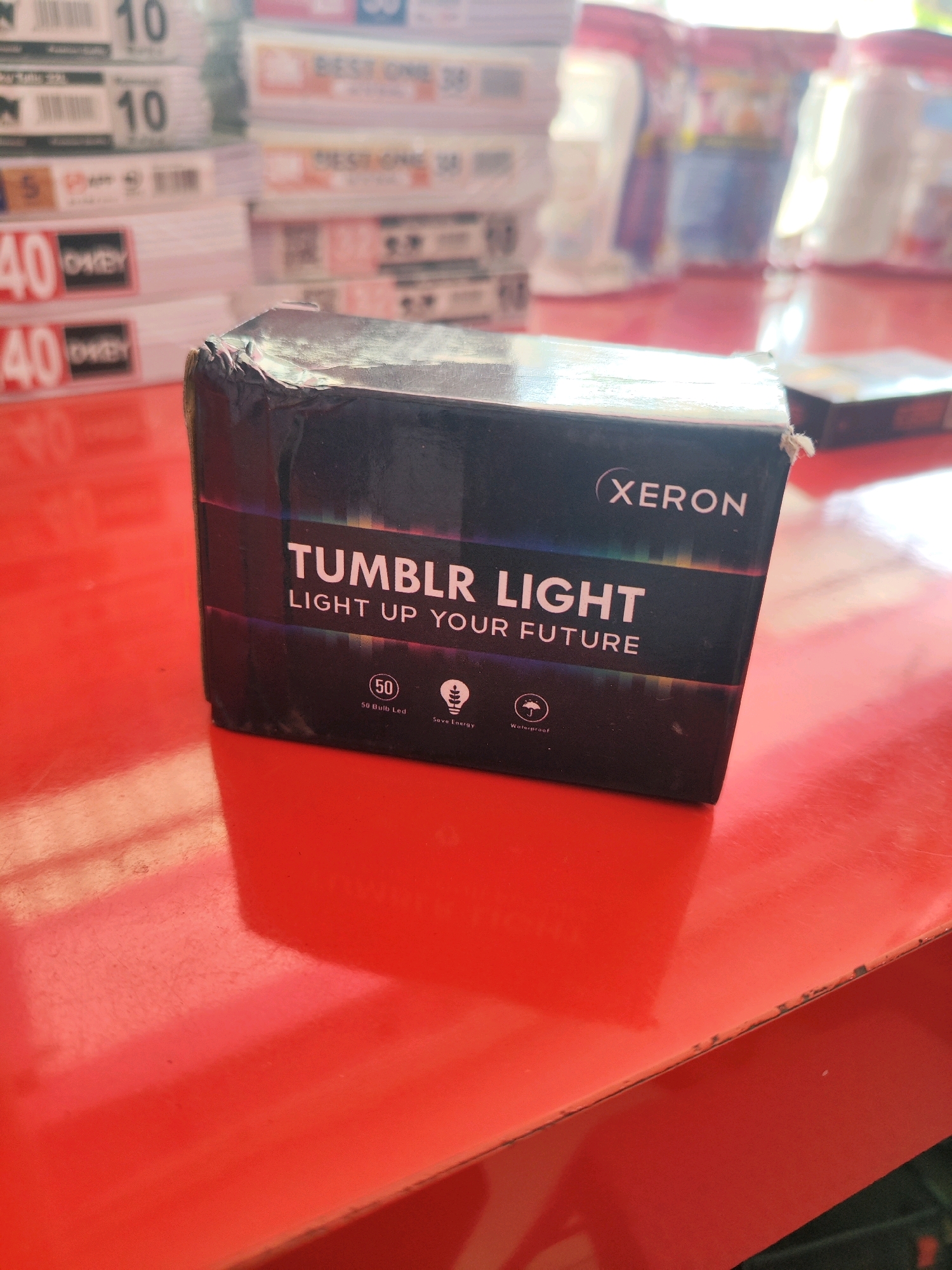 A product box for "Tumblr Light" by "Xeron" sits on a red, reflective surface. The box is black with white text and features a colorful stripe. Below the main text, there are icons indicating "50 Bulb Led," "Save Energy," and "Waterproof." The box appears to be slightly damaged on the corner. In the blurred background, stacks of what appear to be other retail products are visible, some with price tags showing "40" and "10." The overall scene suggests a retail environment, possibly a store in Sragen, Indonesia, given the context.