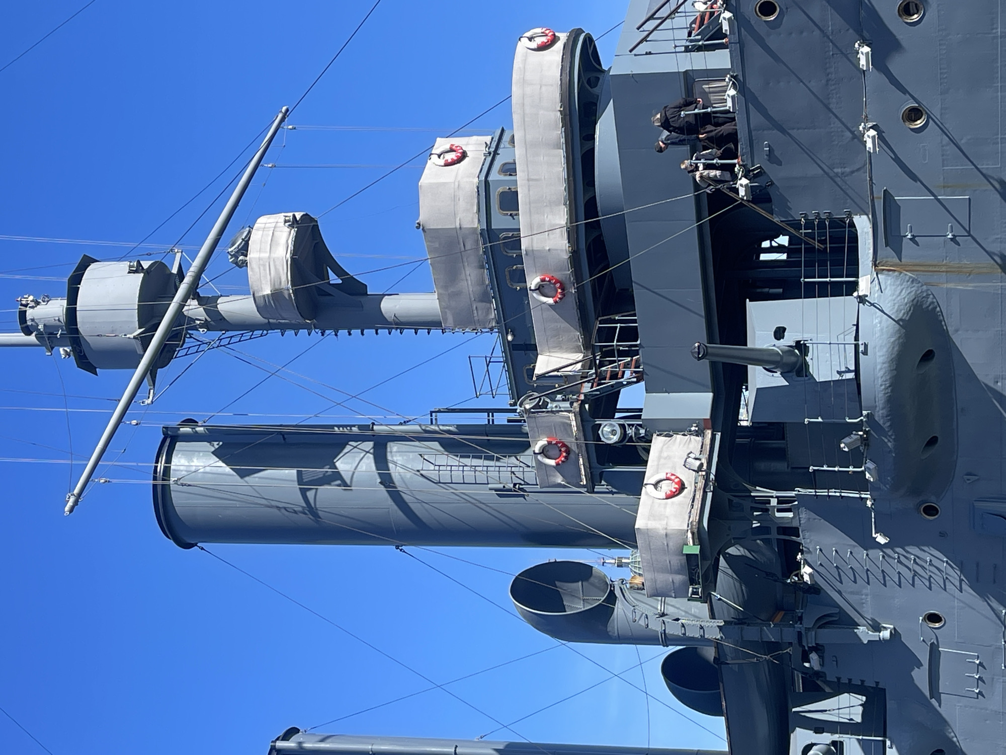 The image shows a portion of the USS Olympia, a retired United States Navy protected cruiser, docked in Philadelphia, Pennsylvania. The view is from a low angle, looking upwards at the ship's superstructure, including its smokestacks, masts, and various platforms. The sky is clear blue. There are no people discernible on the portion of the ship visible from the ground. Life buoys are attached. A gun is visible. The hull has portholes.