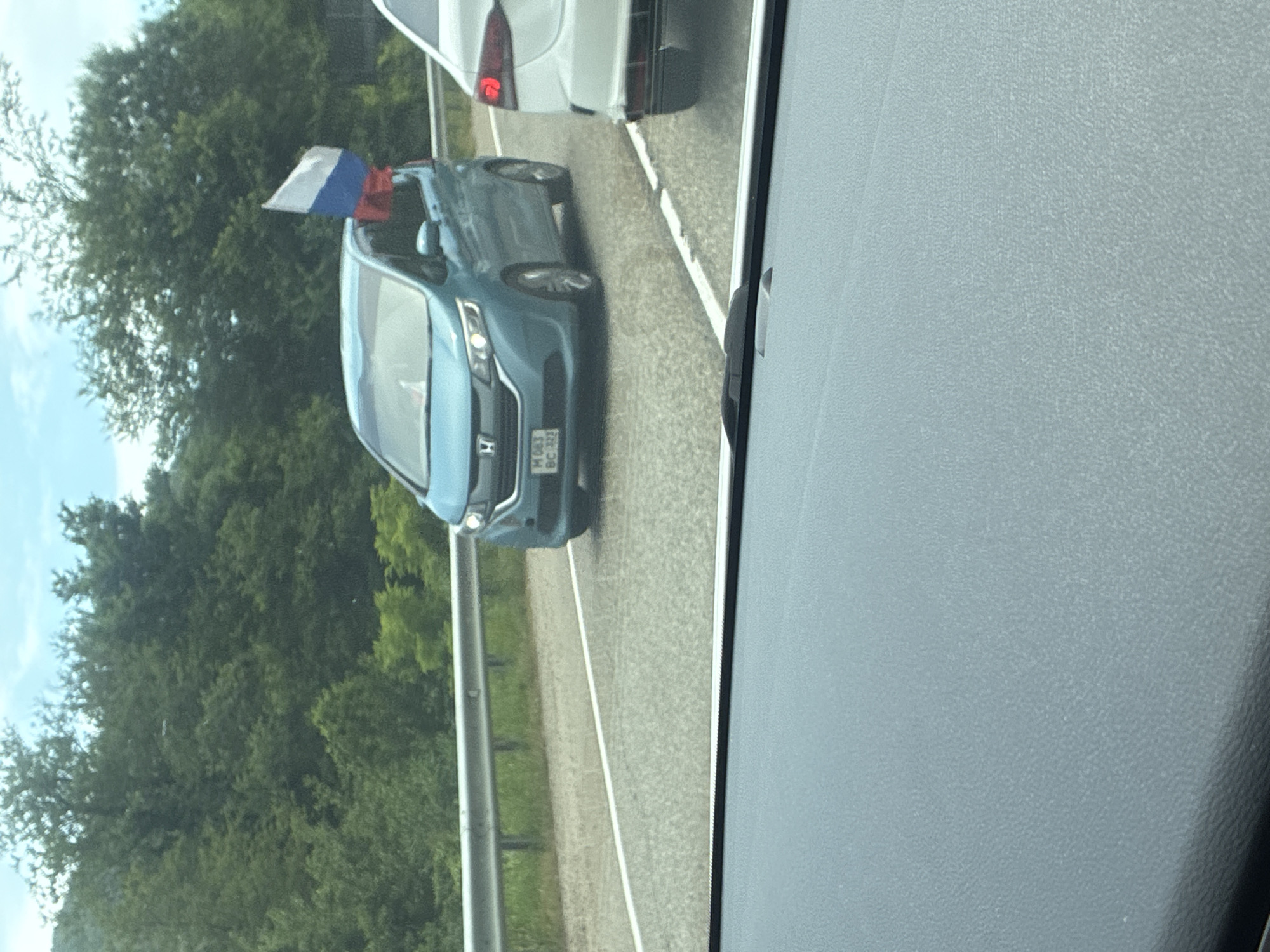 A light blue minivan with a Russian flag attached to its right rear window is driving on a paved road in Arkhipo-Osipovka, Russia. The minivan has a license plate that reads "M 063 BC 717". A white car is driving to the right of the minivan. The road is bordered by a metal guardrail on the left and surrounded by a forest of green trees. The sky is cloudy and blue. The photo appears to be taken from inside another vehicle.