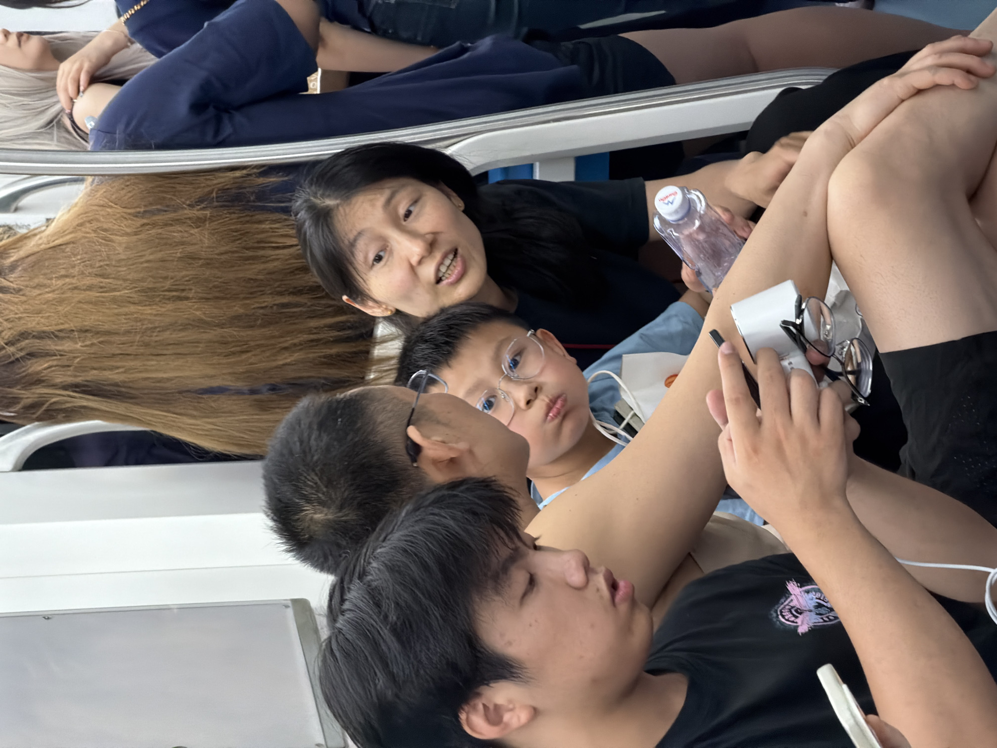 This image depicts the interior of a public transport vehicle, likely a subway car, in the city of Unknown, Unknown. The scene is well-lit and features at least seven individuals of East Asian appearance.
In the foreground, a male youth with short black hair, wearing a black t-shirt with a pink and purple graphic print, is focused downwards on a white smartphone held in his left hand. His right hand manipulates a dark electronic device connected by a white cable. Adjacent and slightly behind him, an adult male with short dark hair and dark-rimmed eyeglasses is partially visible, facing towards the right, with his right arm resting. Next to him, a young boy, approximately 6-10 years old, wearing round spectacles and a light blue t-shirt, makes a puckered mouth expression while looking towards the adult male.
To the boy's right, an adult female with long dark hair, dressed in a black t-shirt, looks towards the left with her mouth slightly agape, holding a clear plastic water bottle with a white and red cap in her right hand. In the background, to the right, a standing individual in a dark blue t-shirt and dark shorts is partially visible from the waist down. Further in the upper background, another individual with long blonde/grey hair is seen, along with a person displaying long light brown hair on the upper left. The vehicle's interior features white panels and prominent silver metal grab rails.