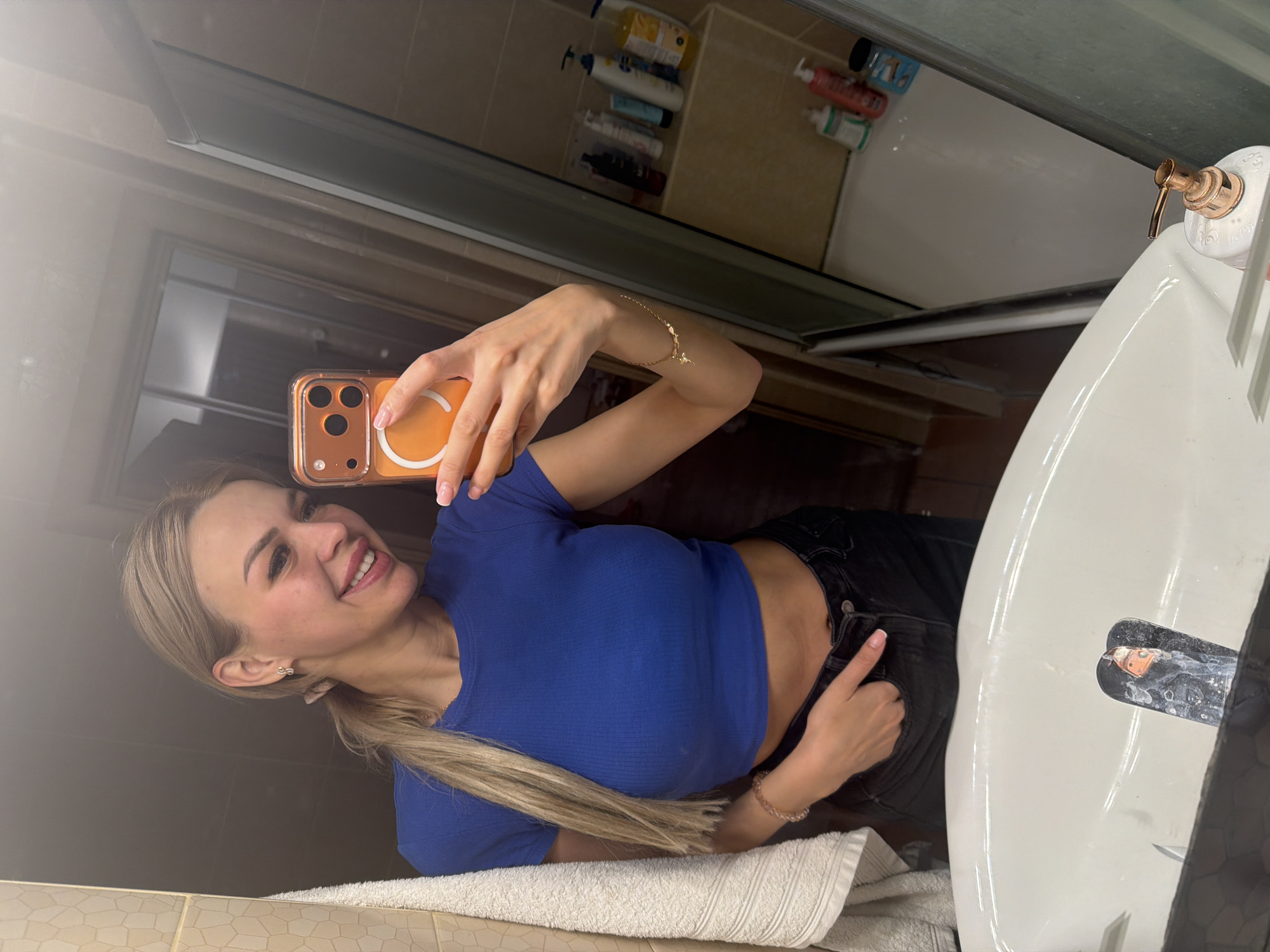 A woman is taking a mirror selfie in a bathroom. She has blonde hair pulled back in a ponytail and is wearing a blue crop top and black jeans. She is holding an orange phone with a white ring light attachment. The bathroom appears to be in a residential setting, with a sink, mirror, and shower visible. There are various toiletries on a shelf in the background. The location is the city of Kyiv, Ukraine.