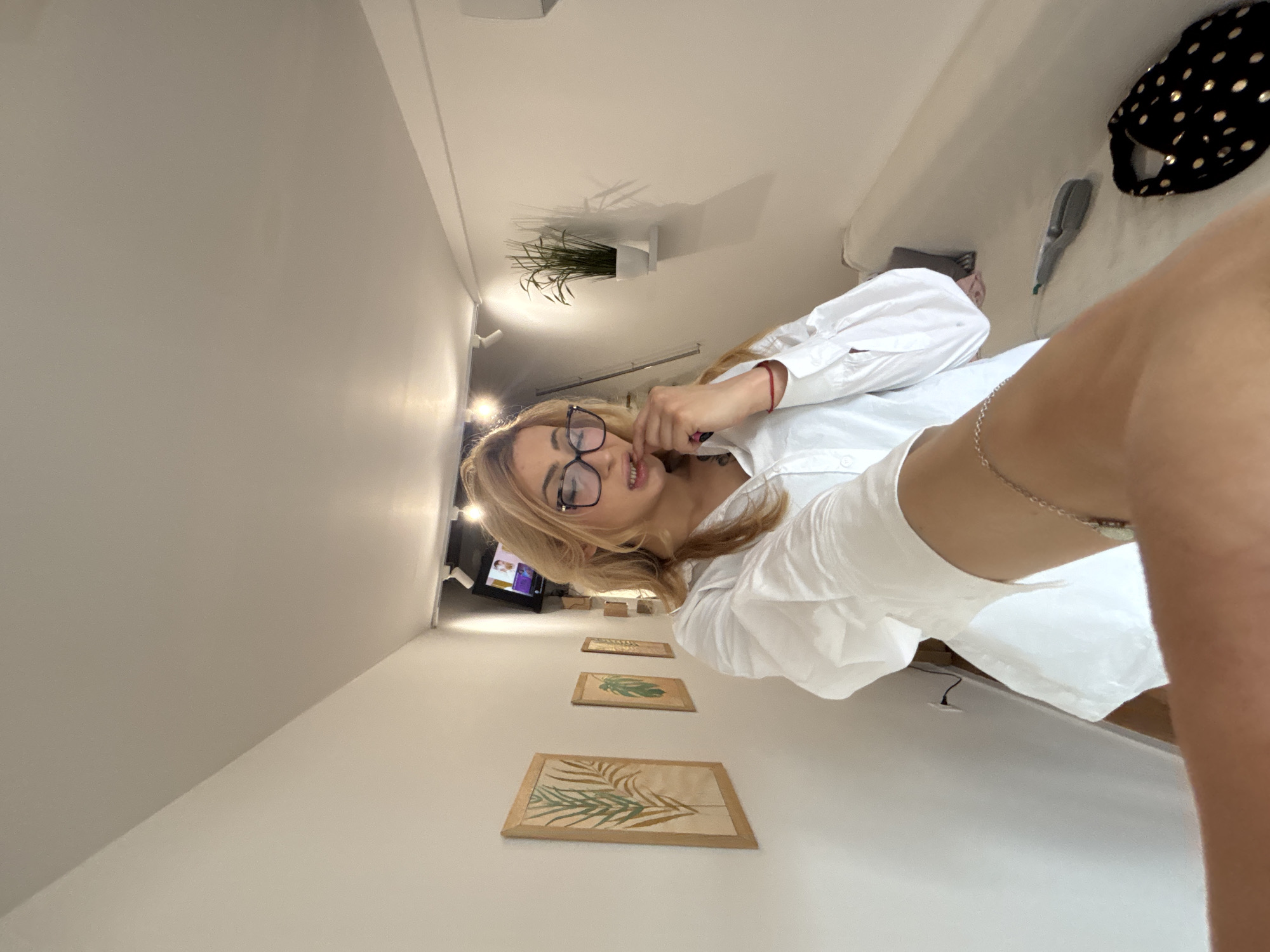 A waist-up shot depicts a fair-skinned woman with blonde hair, wearing glasses and a white button-down shirt. She is indoors, presumably in a building in Varna, Bulgaria. Behind her, wall art featuring botanical prints hangs. A houseplant sits on a high shelf, and a black bag is near a hair dryer. The scene appears to be a living or dressing space.