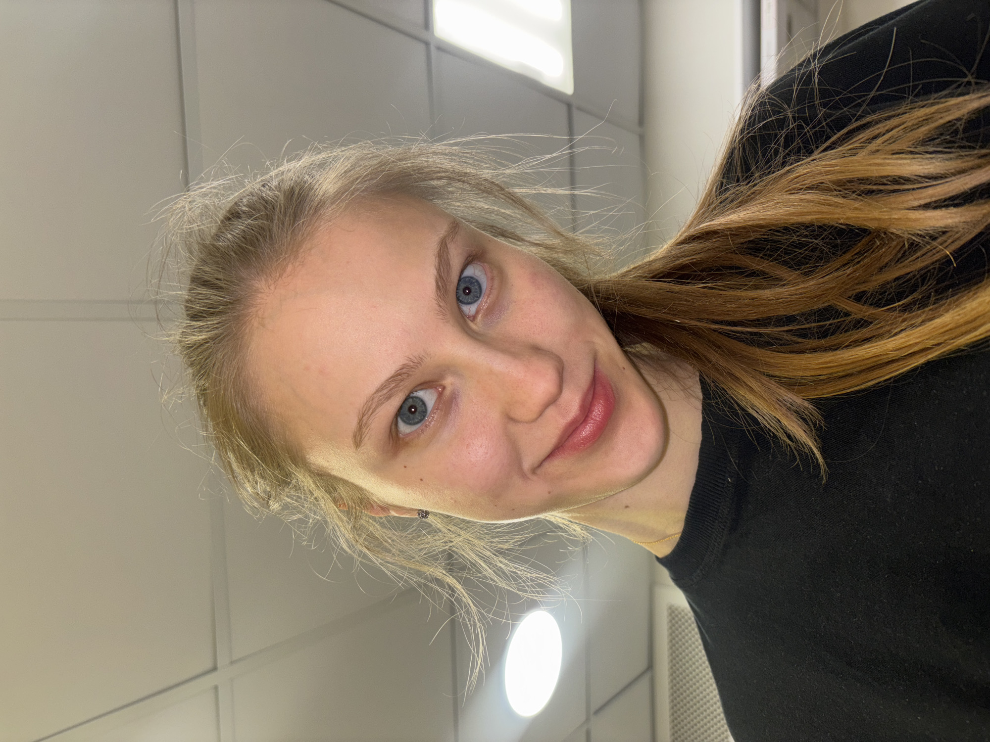 A young woman with fair skin, blue eyes, and blonde-brown hair is depicted in a selfie. She is wearing a black top. The background consists of a white paneled wall and ceiling lights. The location in Perm, Russia is not apparent from the image.
