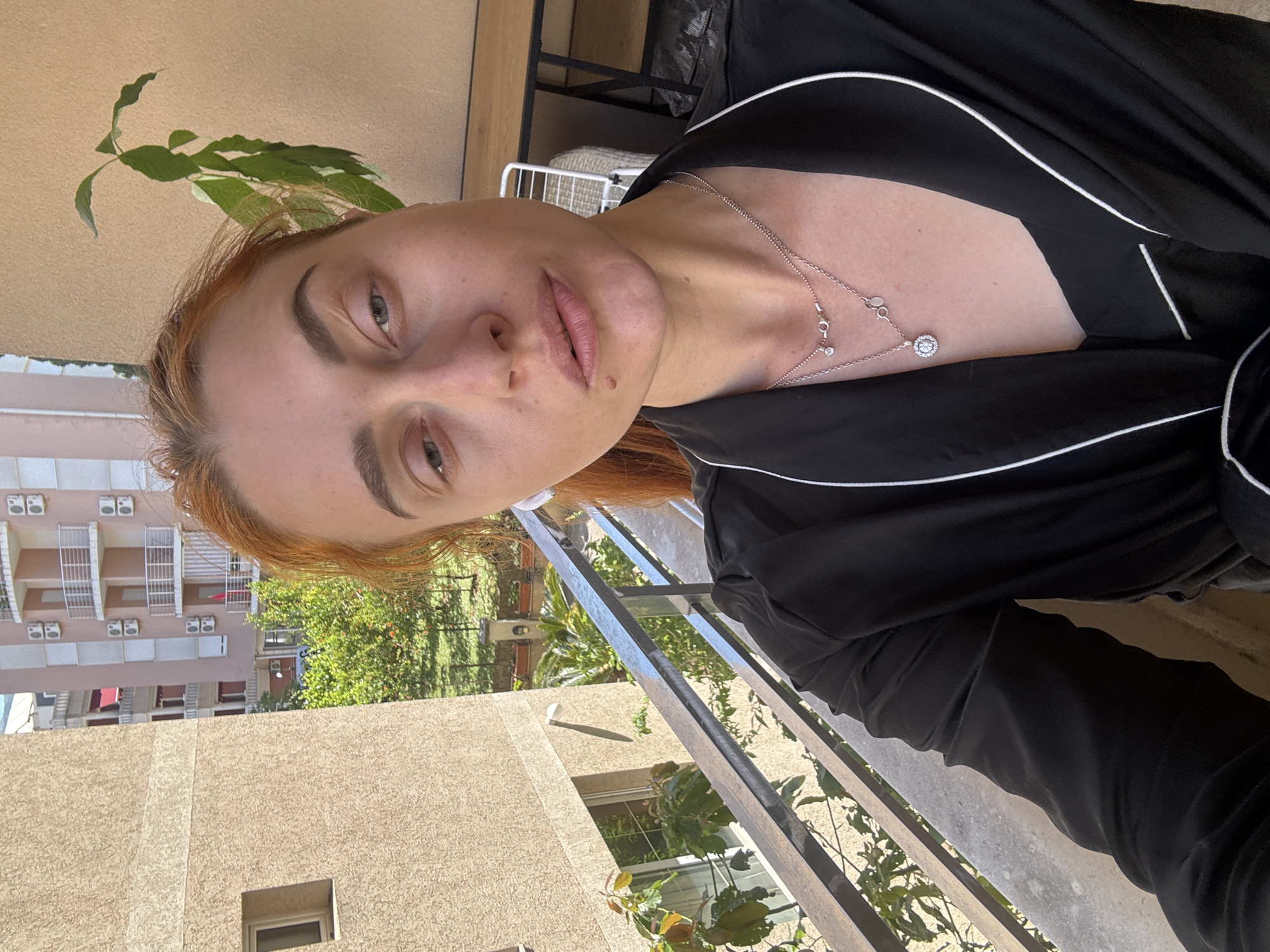 A woman with reddish-blonde hair is photographed from a high angle on a balcony. She is wearing a black pajama-style top with white piping and a silver necklace. The background includes a beige building with multiple windows and air conditioning units, a railing of the balcony, and greenery outside. The location is Budva, Montenegro.