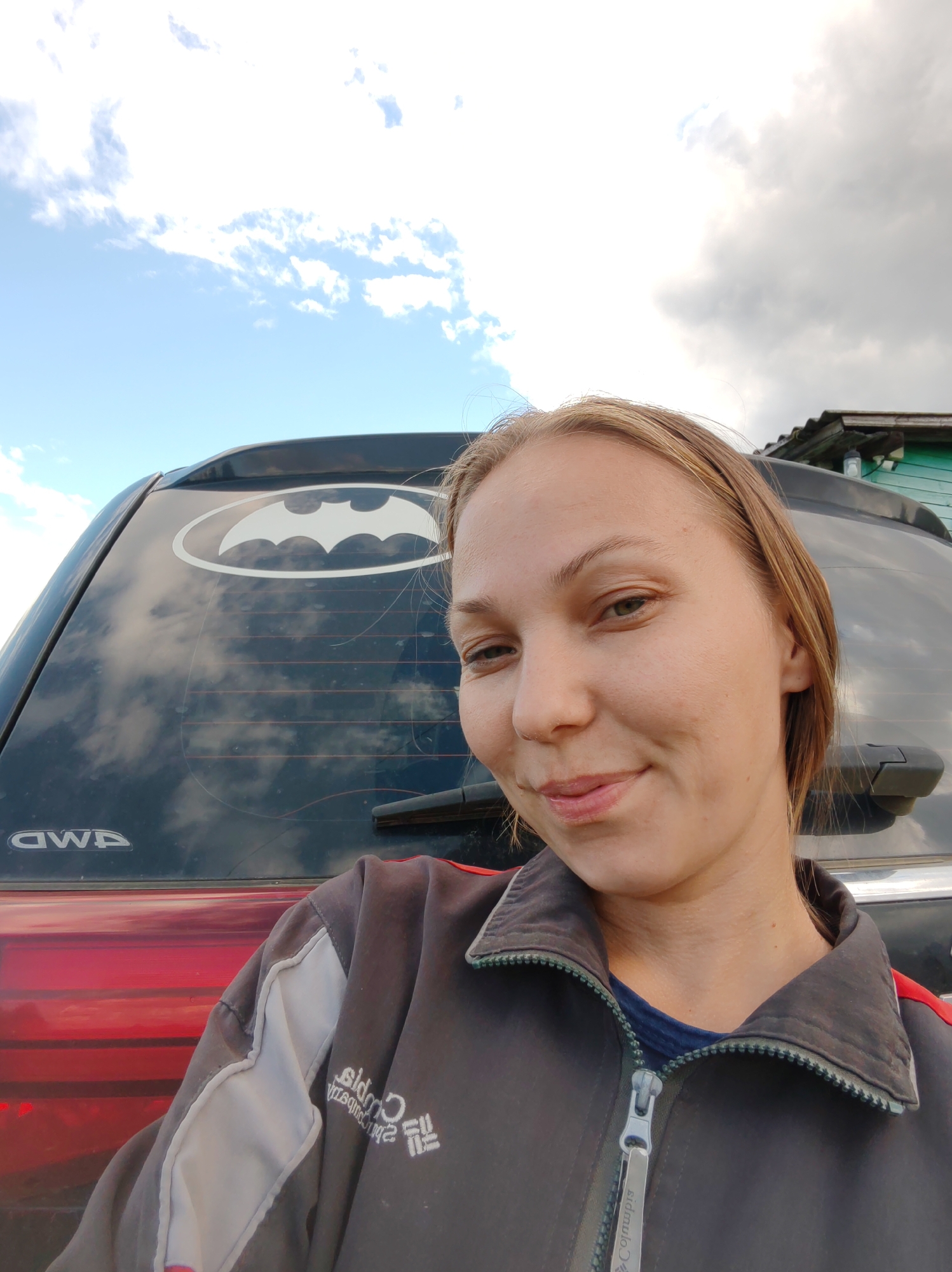 In an outdoor, low-angle, selfie-style shot, a young woman with light brown hair and green eyes smiles at the camera. She is wearing a dark gray jacket with white and red accents over a dark blue shirt. The jacket has a zipper and embroidered logos. Behind her, the rear of a dark-colored SUV is visible, featuring a white Batman logo sticker on the rear window and "4WD" branding near the bottom. The SUV's window reflects the sky and clouds. To the right of the SUV, a portion of a green wooden building with a corrugated roof is visible. The environment appears to be a rural or suburban setting, possibly the city of Chukhlem, Russia, indicated by the presence of the building and the overcast sky with patches of blue.