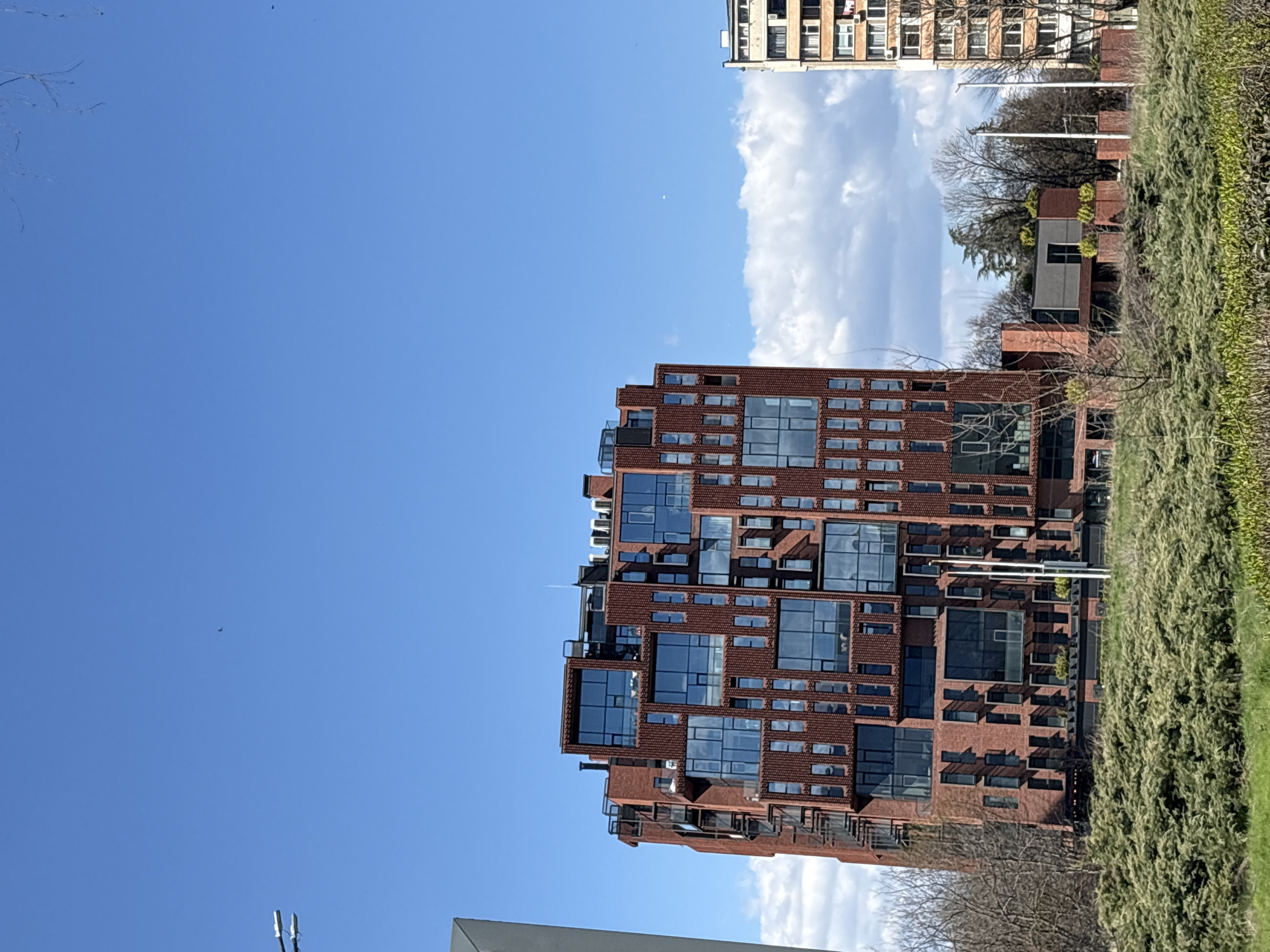 The image shows a modern brick building with numerous windows in Sofia, Bulgaria, set against a clear blue sky with some clouds. Adjacent to the building is a green space with vegetation. In the background, another, older building is partially visible. There are no people visible in the scene. The image appears to be taken during the daytime.