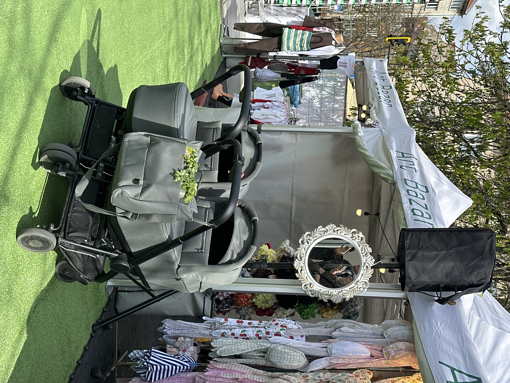 The image shows an outdoor scene at an "Art Bazar" market stall, likely in Sofia, Bulgaria. The stall is set up under a white tent-like structure with "Art Bazar" written in green. In front of the stall is a double stroller with a gray body and black frame. A bag is attached to the stroller with some greenery. Inside the stall, a shelf displays handmade items, including patterned fabrics, scrunchies, and a decorative mirror with a person reflected in it. A black bag or container sits on a stand next to the mirror. A clothes rack with hanging garments is visible in the background. The ground is covered with artificial grass. The background reveals a cityscape, including buildings and trees. A traffic light is visible in the distance.