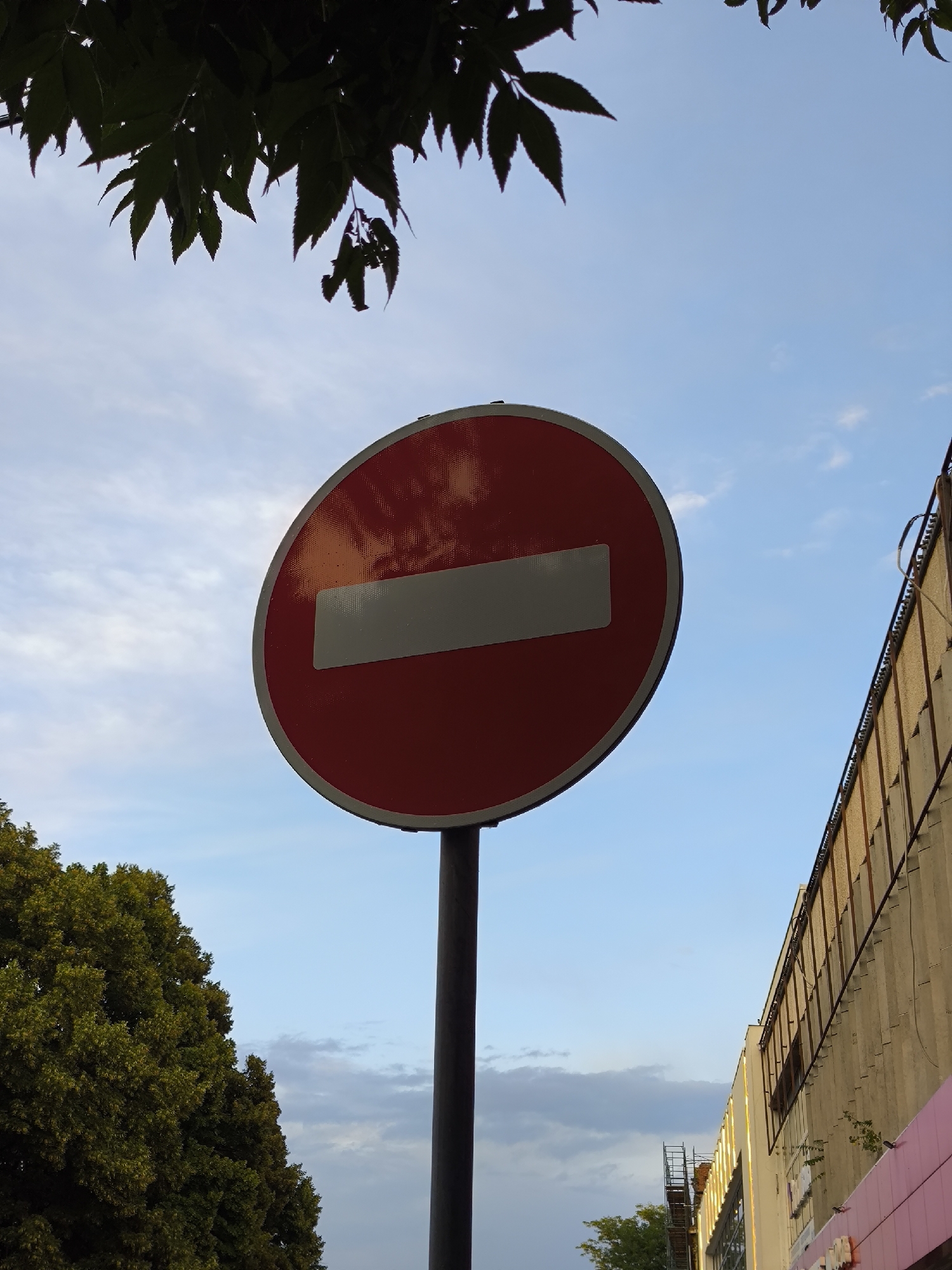 An outdoor, low-angle, medium shot captures a red and white "Do Not Enter" sign against a partly cloudy blue sky. The circular sign, with a horizontal white bar in the center, is mounted on a dark pole. To the left of the sign, the dark green foliage of a tree is visible, partially obscuring the sky. To the right, the textured concrete facade of a building, lined with metal grates and a pink trim, rises diagonally. The lighting suggests it is daytime. The scene appears to be a street or sidewalk area in Yessentuki, Russia.